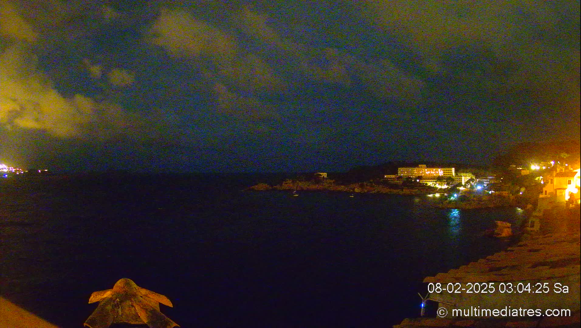 A nighttime coastal scene shows a dark body of water with dimly lit buildings across the water under a partly cloudy sky.