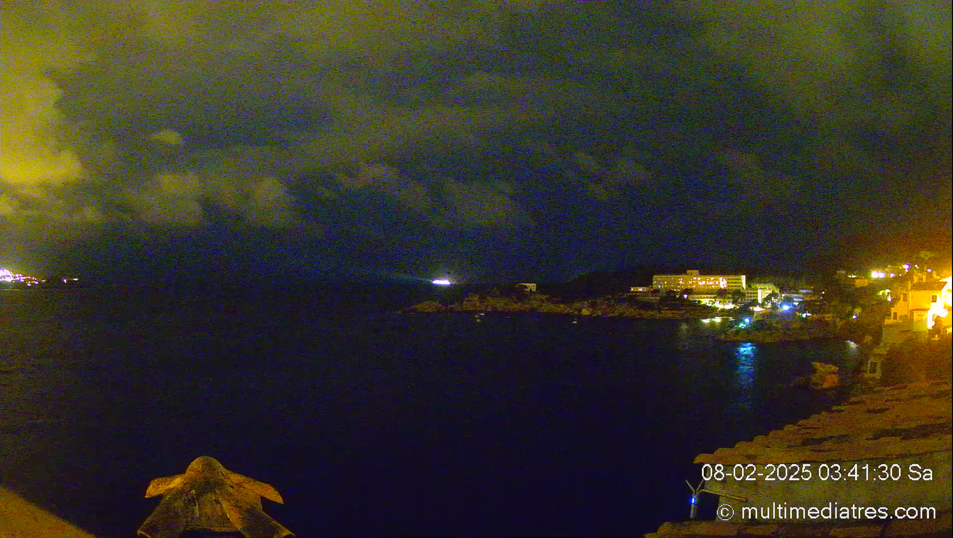 A nighttime coastal scene shows a dark body of water under a cloudy sky, with illuminated buildings visible on the shore.