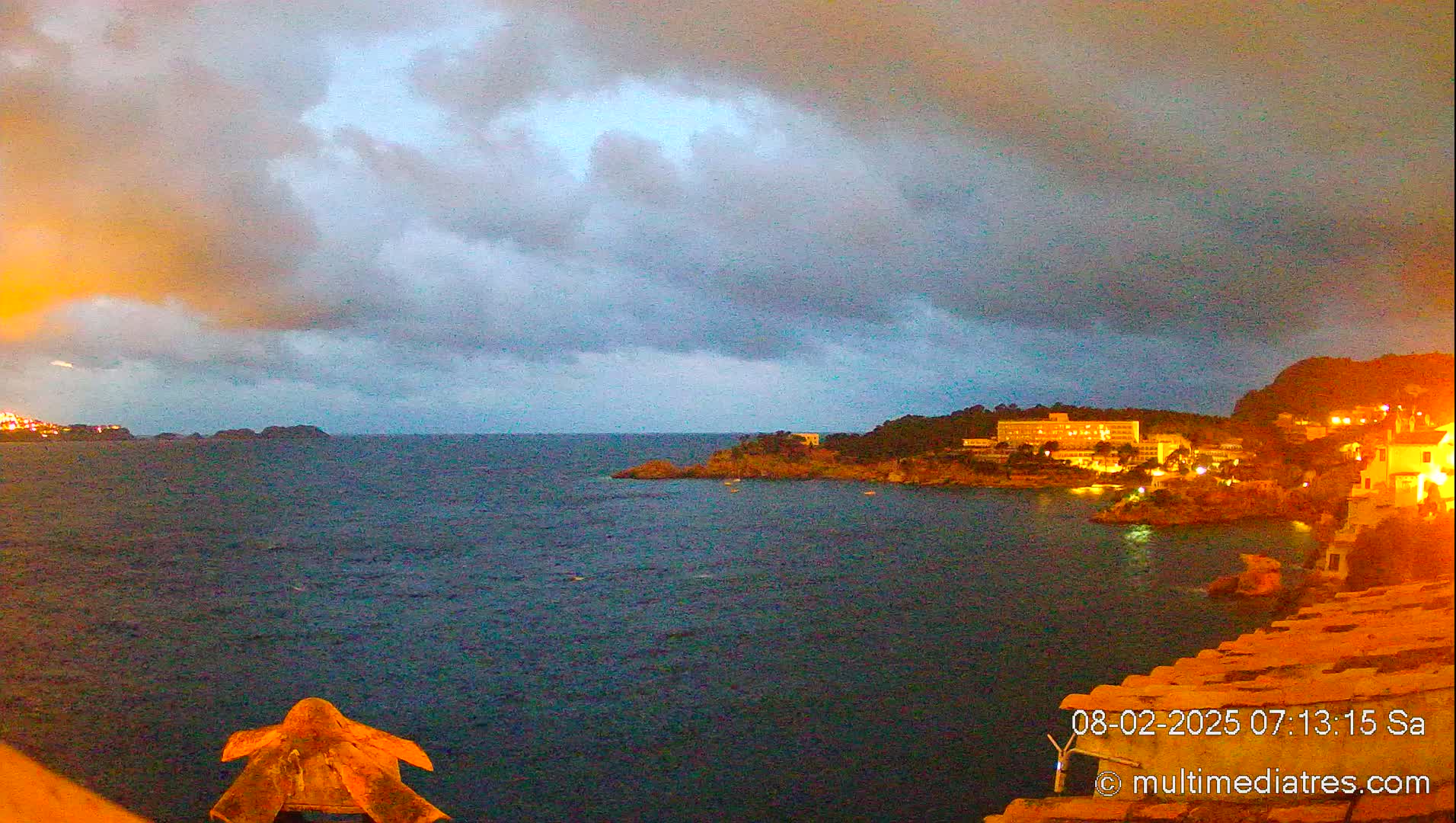 A partly cloudy, dusky evening scene overlooks a dark-blue ocean, with a coastline featuring buildings lit up at night.