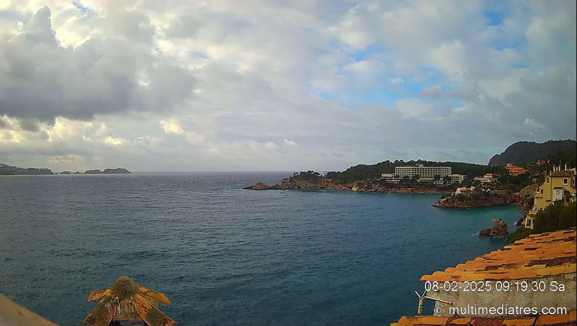 A partly cloudy day shows a coastal view of a calm sea with distant islands, a hotel, and buildings nestled along a rocky coastline.