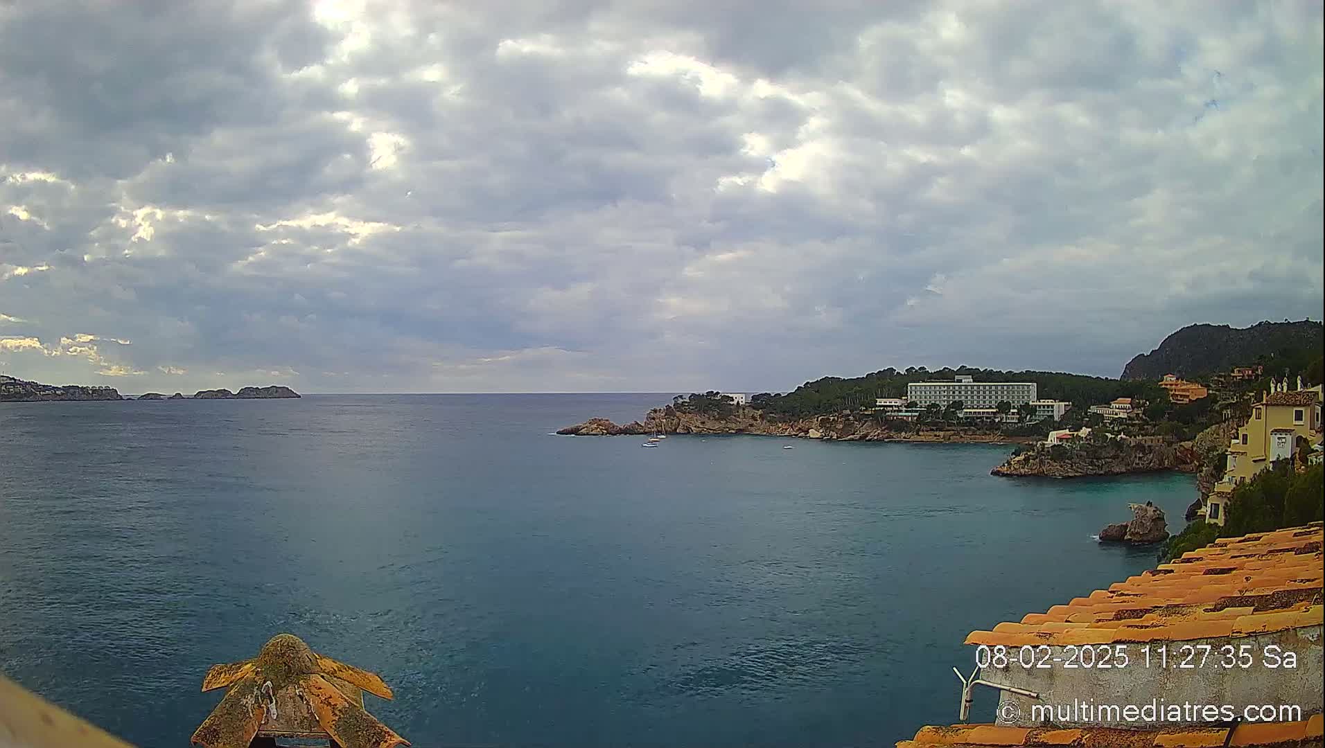A partly cloudy day overlooks a calm bay with a hotel and buildings along the rocky coastline.