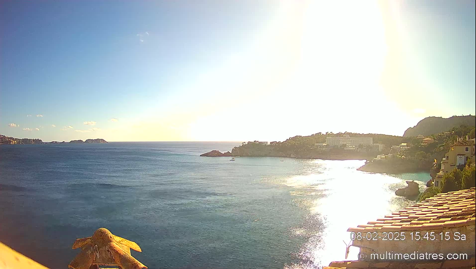 A sunny coastal scene shows a calm sea with small islands in the distance, a rocky shoreline with buildings, and a tiled roof in the foreground.