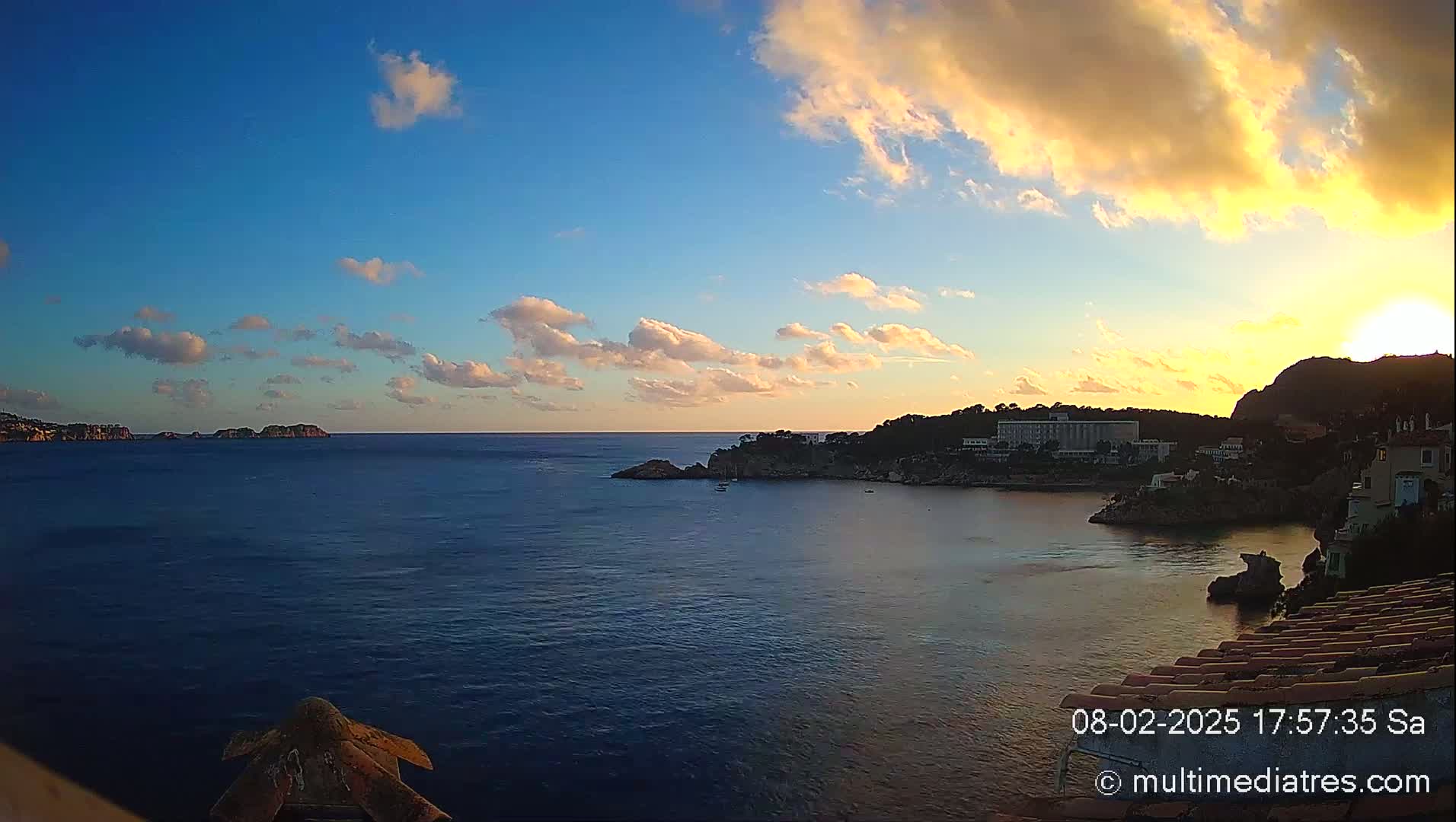 A coastal sunset scene shows a calm body of water with a town nestled on a rocky shoreline under a partly cloudy, golden sky.