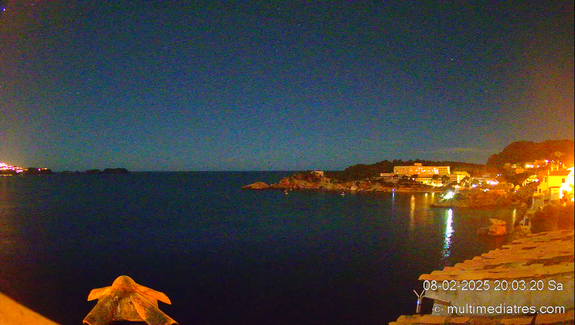 A nighttime view of a dark-blue ocean bay, with a coastline featuring illuminated buildings and a clear, starry sky.