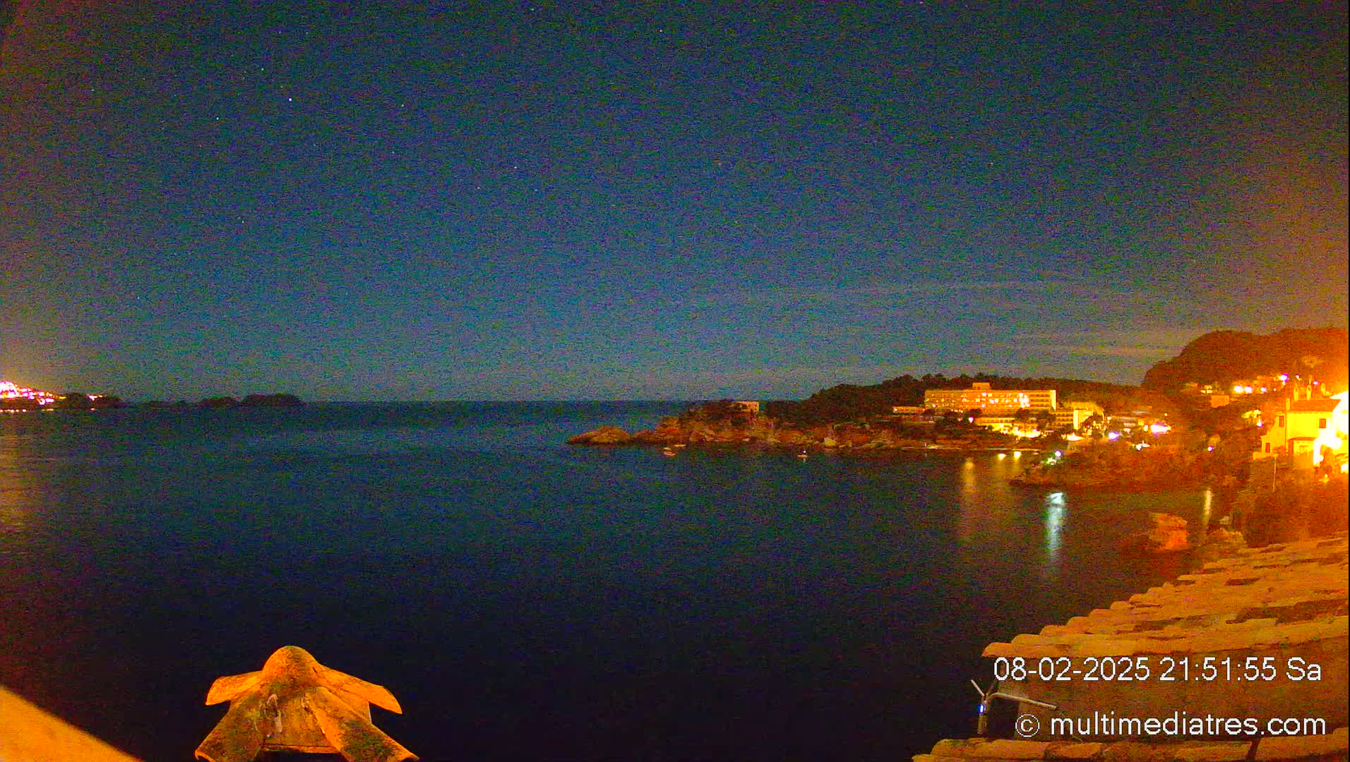 A nighttime view of a calm, dark-blue sea with a dimly lit coastal town on a hill under a clear, starry sky.