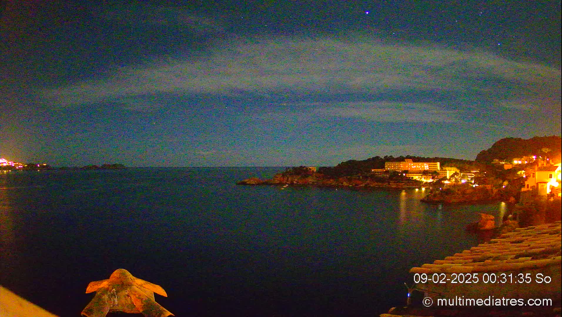 A nighttime coastal scene shows a dark body of water with illuminated buildings on a distant shore under a mostly clear, starry sky.