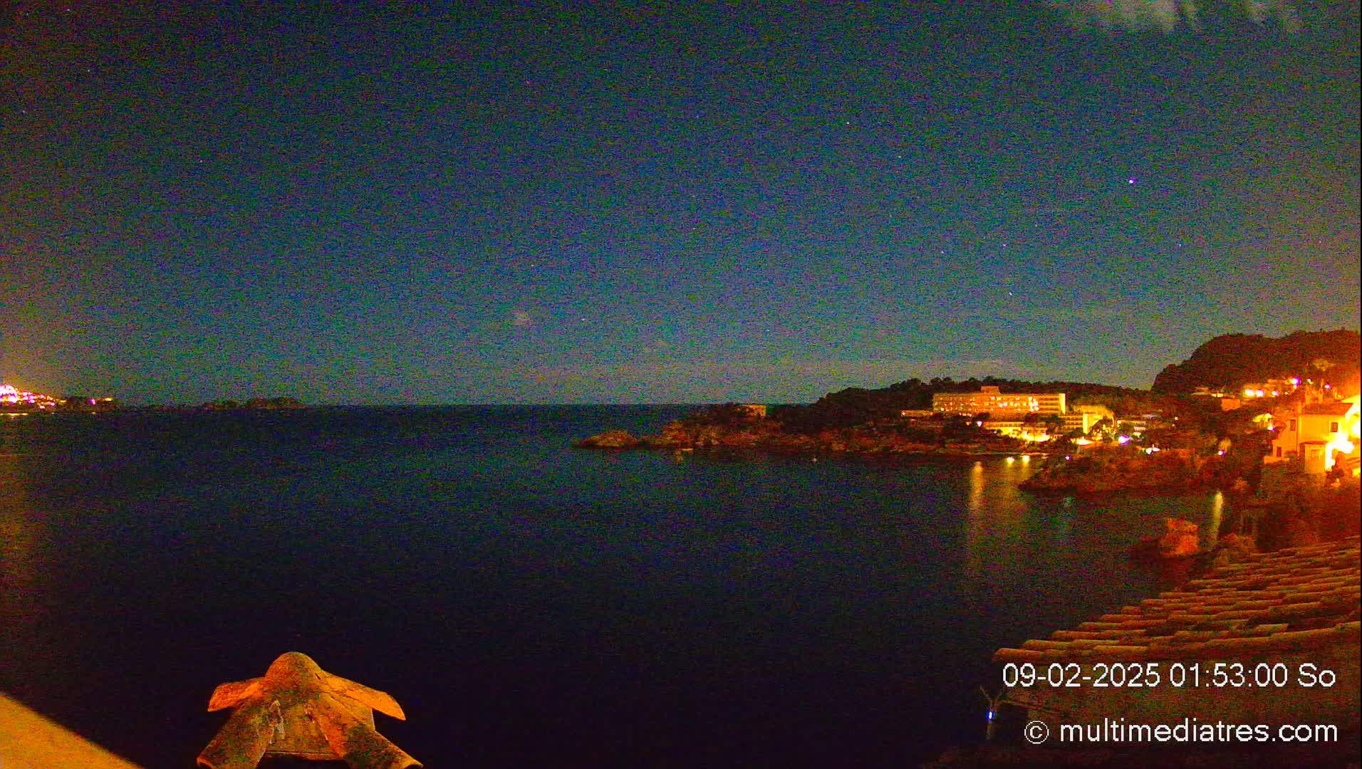 A nighttime coastal scene shows a dark body of water reflecting the lights of buildings on the shore under a clear, starlit sky.