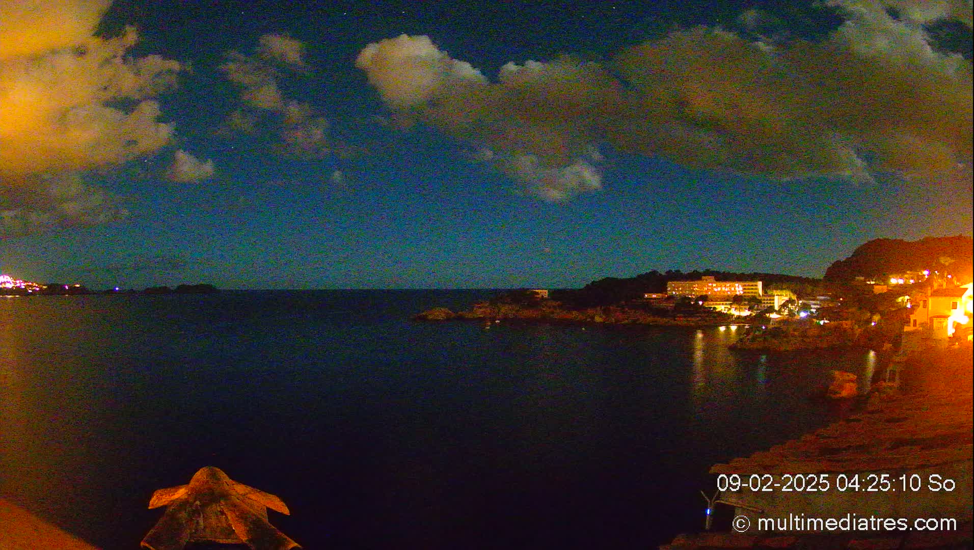 A nighttime coastal scene under a partly cloudy sky shows a dark body of water with illuminated buildings on the opposite shore.