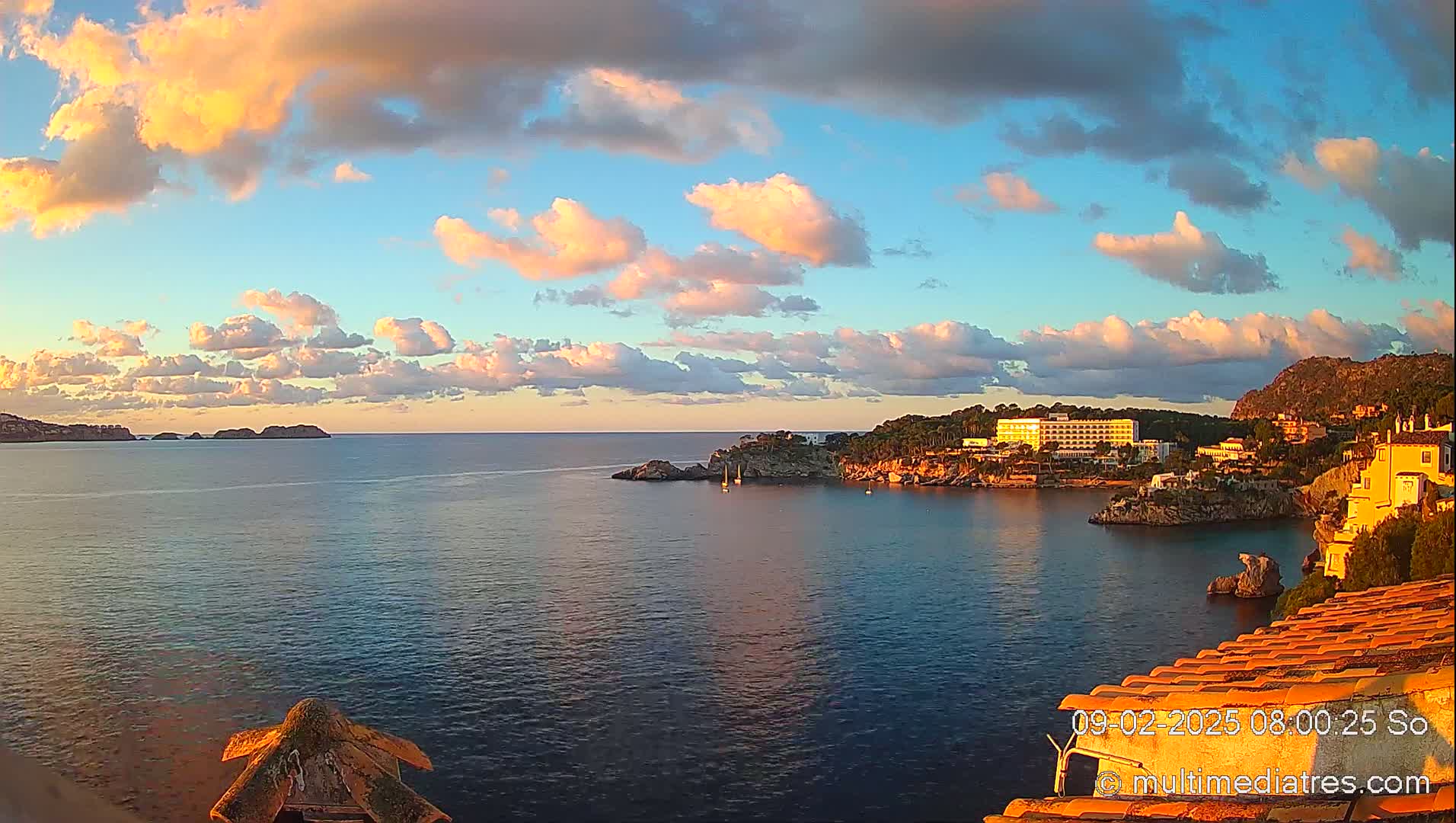 A coastal scene at sunset shows a calm sea with a hotel and buildings nestled along a rocky shoreline under a partly cloudy sky.