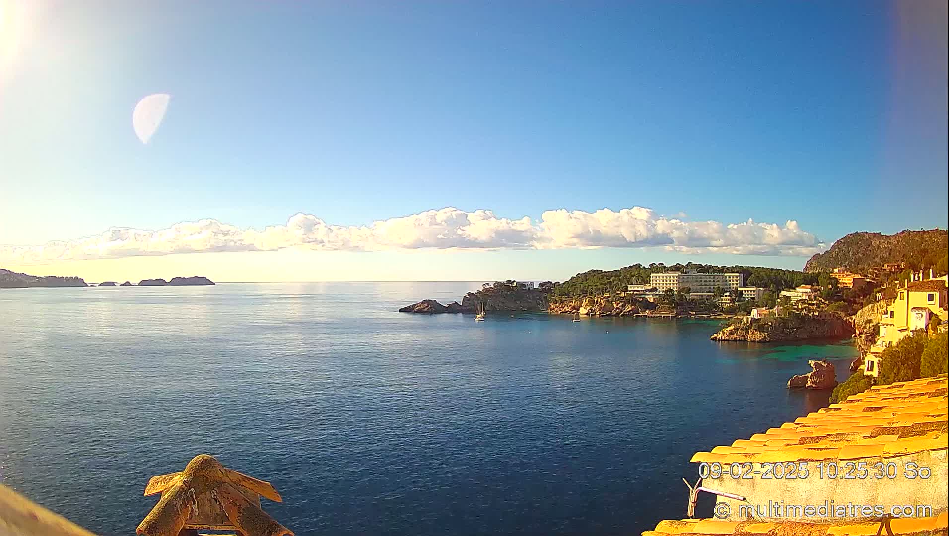 A partly cloudy sunny day shows a coastal scene with a calm blue sea, a crescent moon in the sky, and buildings situated on a rocky hillside overlooking the water.