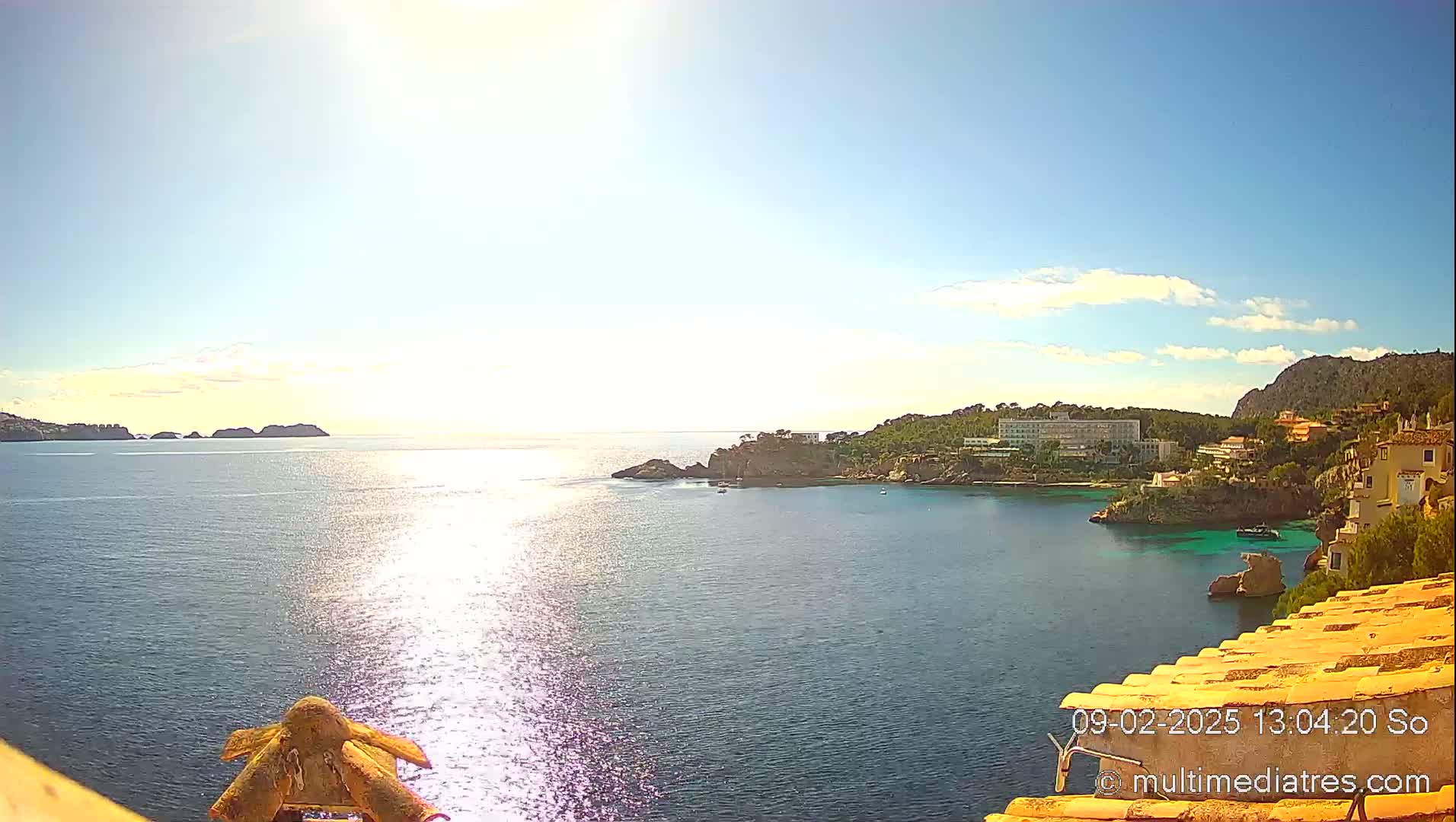A sunny day reveals a coastal view of a calm bay with rocky shores, buildings nestled into the hillside, and small islands in the distance.