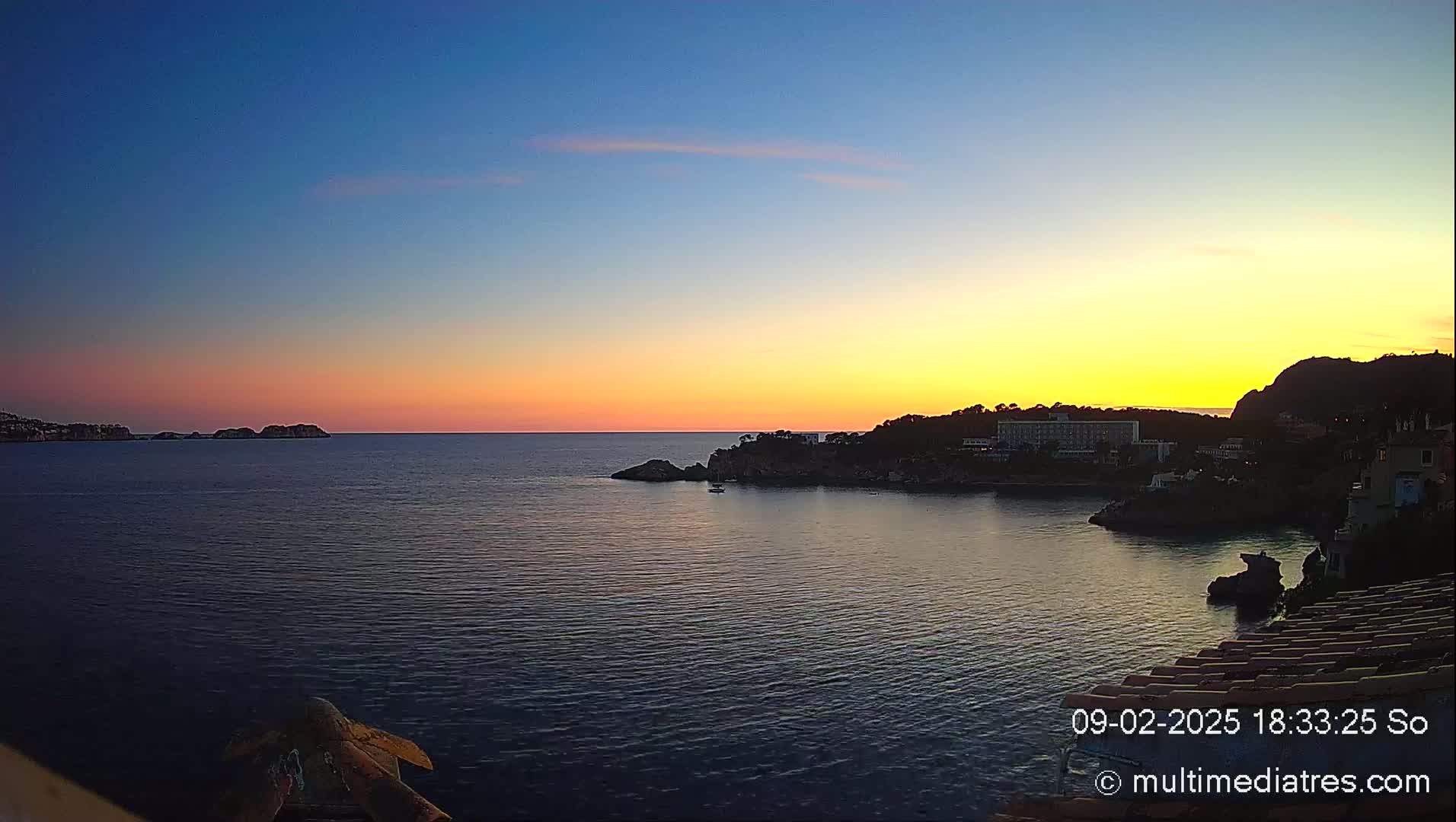 A coastal sunset over calm water shows a town nestled on a rocky shoreline with islands in the distance.