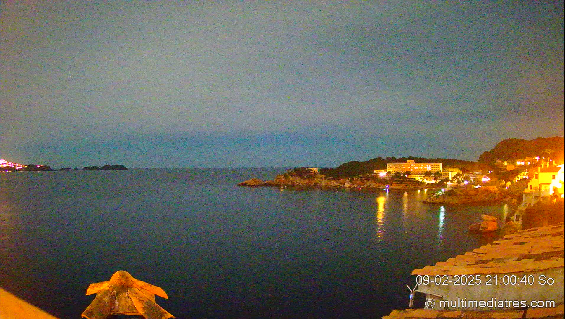 A nighttime coastal scene shows a calm body of water reflecting the lights of buildings on a nearby shore under a dark, clear sky.