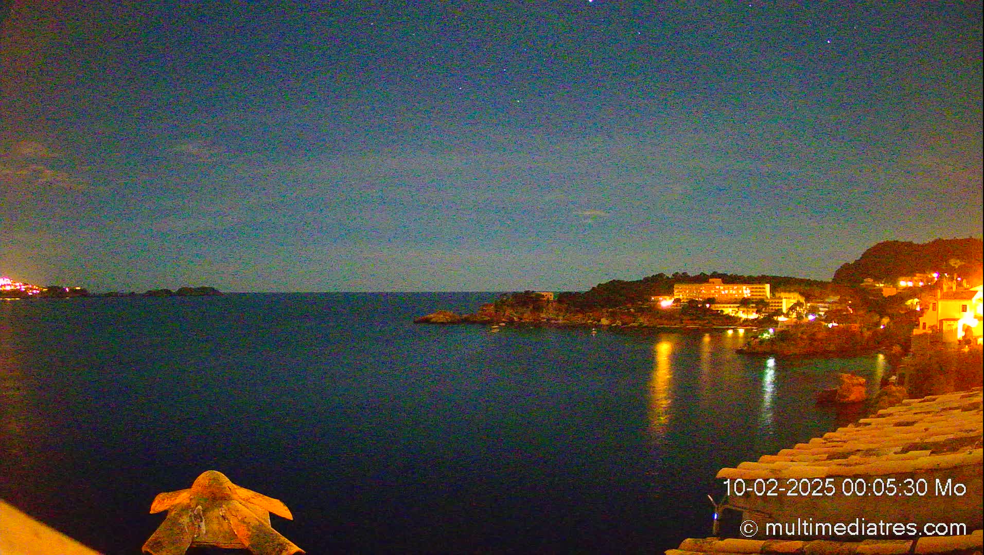A nighttime view of a calm, dark-blue sea reflecting the lights of buildings on a distant shore under a clear, starry sky.