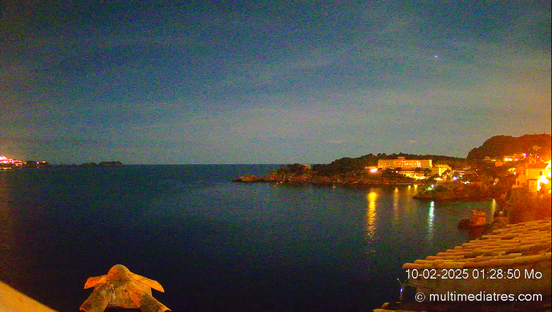 A nighttime view of a calm body of water with illuminated buildings on the opposite shore under a clear, dark sky.