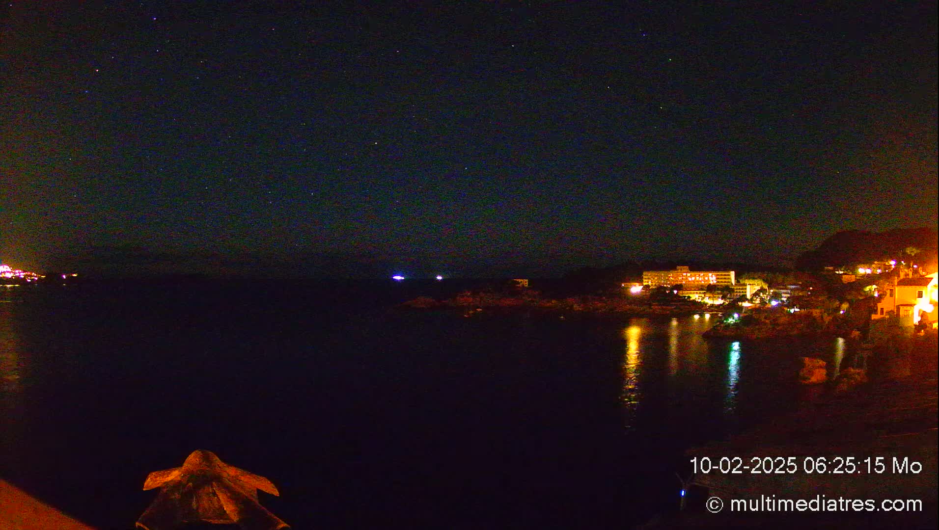 A nighttime coastal scene shows a dark body of water reflecting the lights of buildings on a hillside under a starry sky.