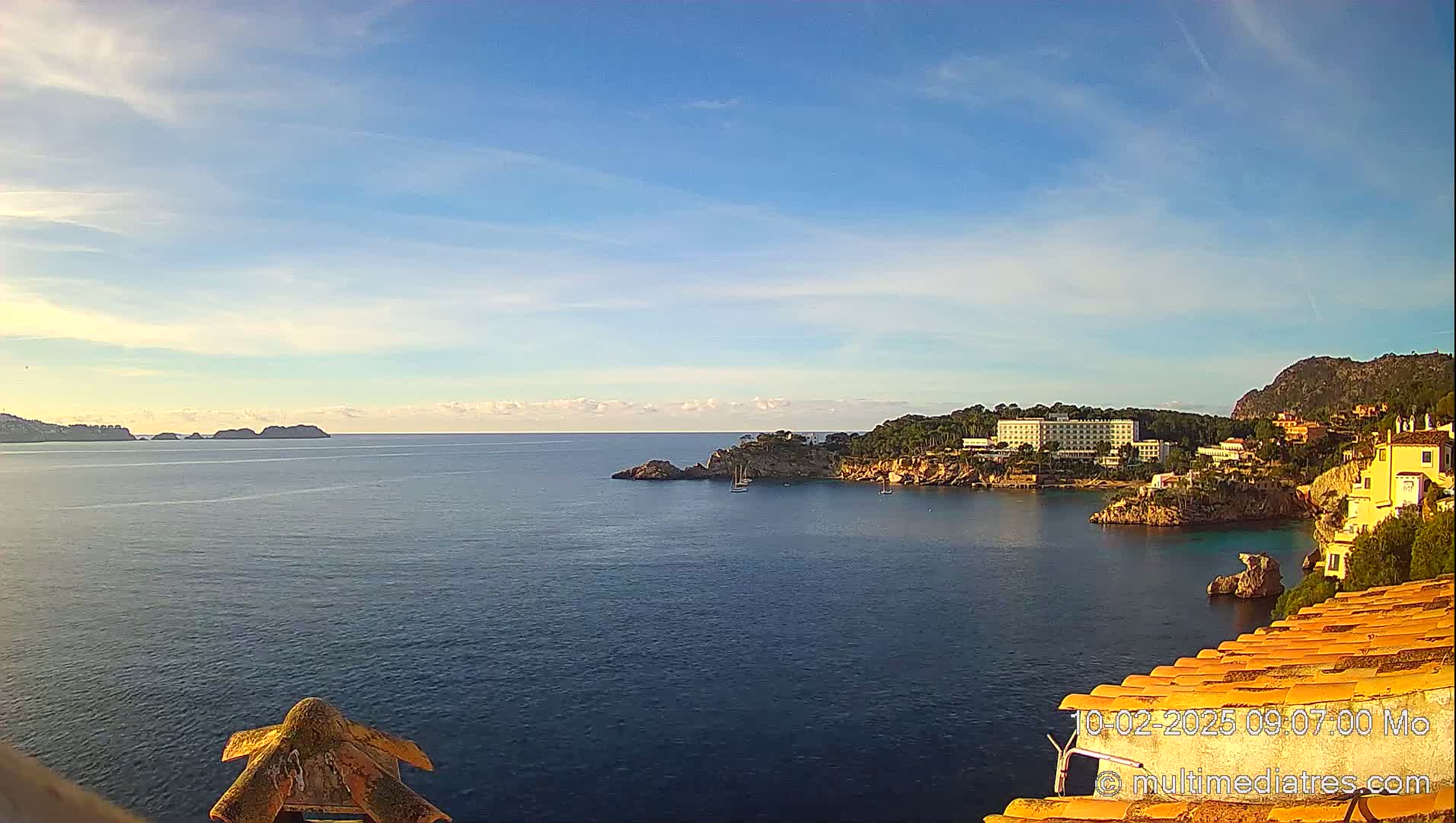 A calm sea with a coastal town and hotel nestled into a rocky cove under a mostly clear, bright sky.