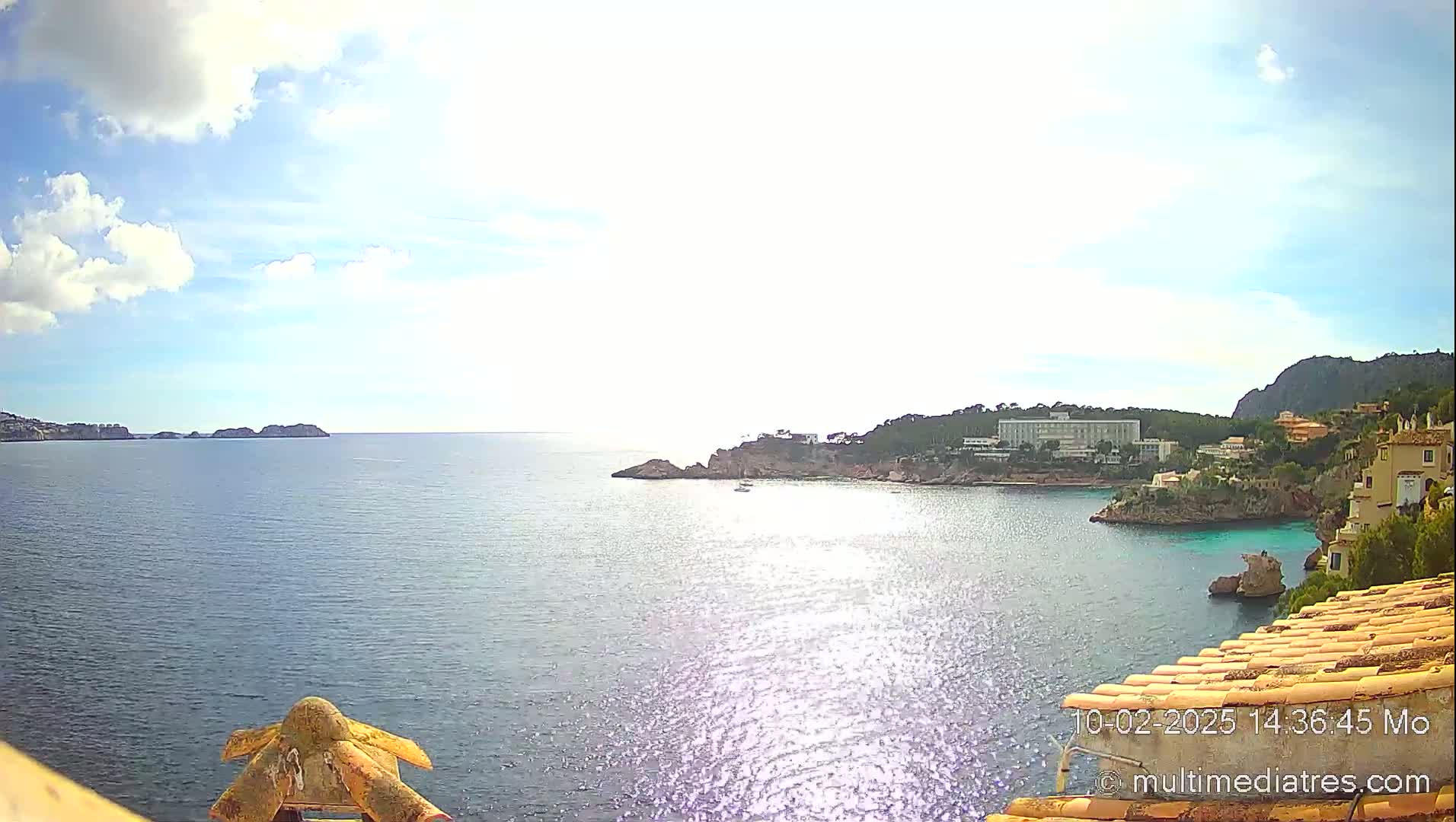 A sunny, partly cloudy day overlooks a calm bay with a coastline of rocky cliffs and buildings, including a large white hotel.