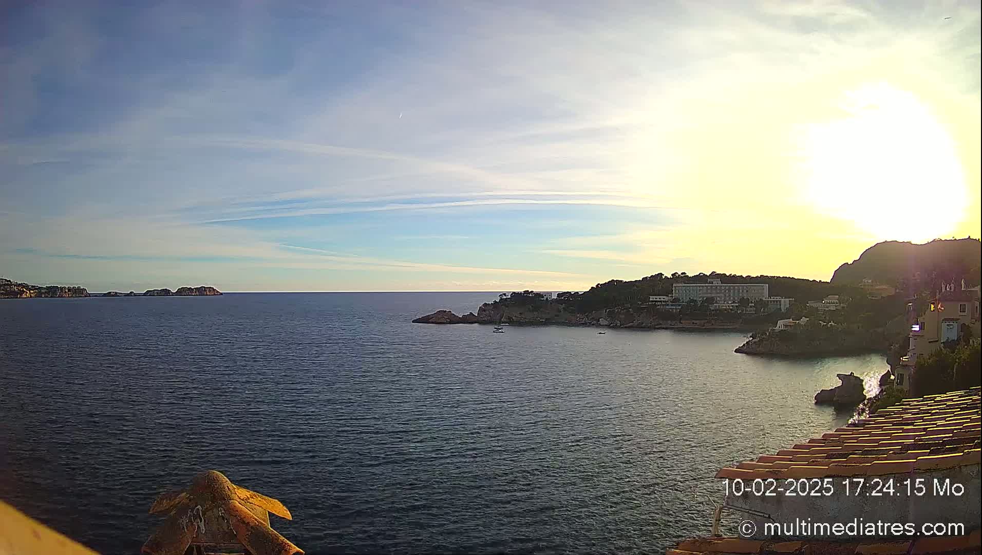 A calm sea with a coastline featuring buildings and rocky outcrops is seen under a mostly sunny sky.