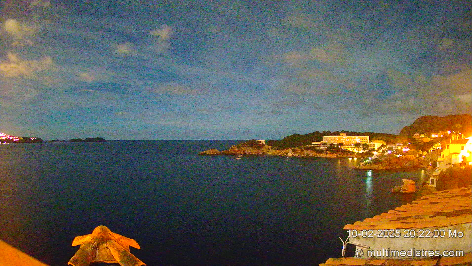 A nighttime coastal scene shows a dark blue sea under a mostly clear sky with a few clouds, with illuminated buildings and vegetation along the shoreline.