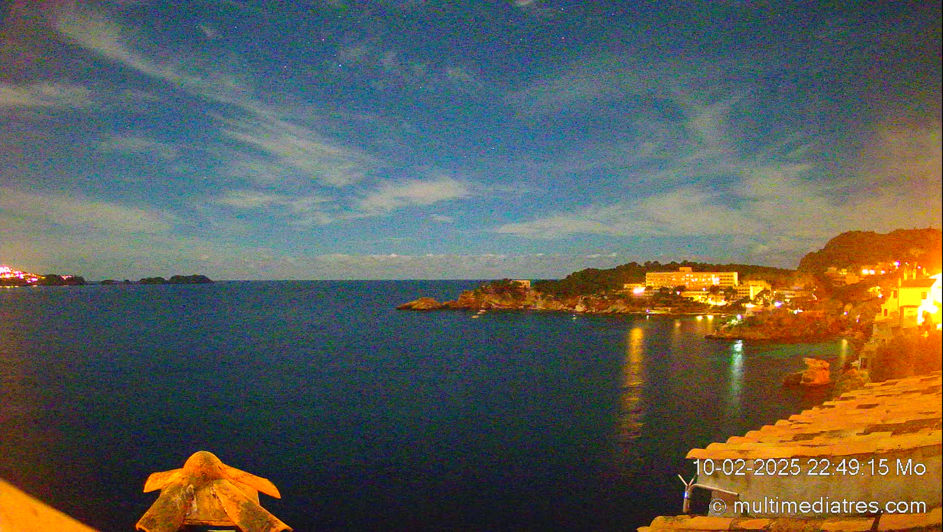 A nighttime coastal scene shows a dark blue sea under a mostly clear, starry sky with thin clouds, illuminated by the lights of buildings on a distant shore.