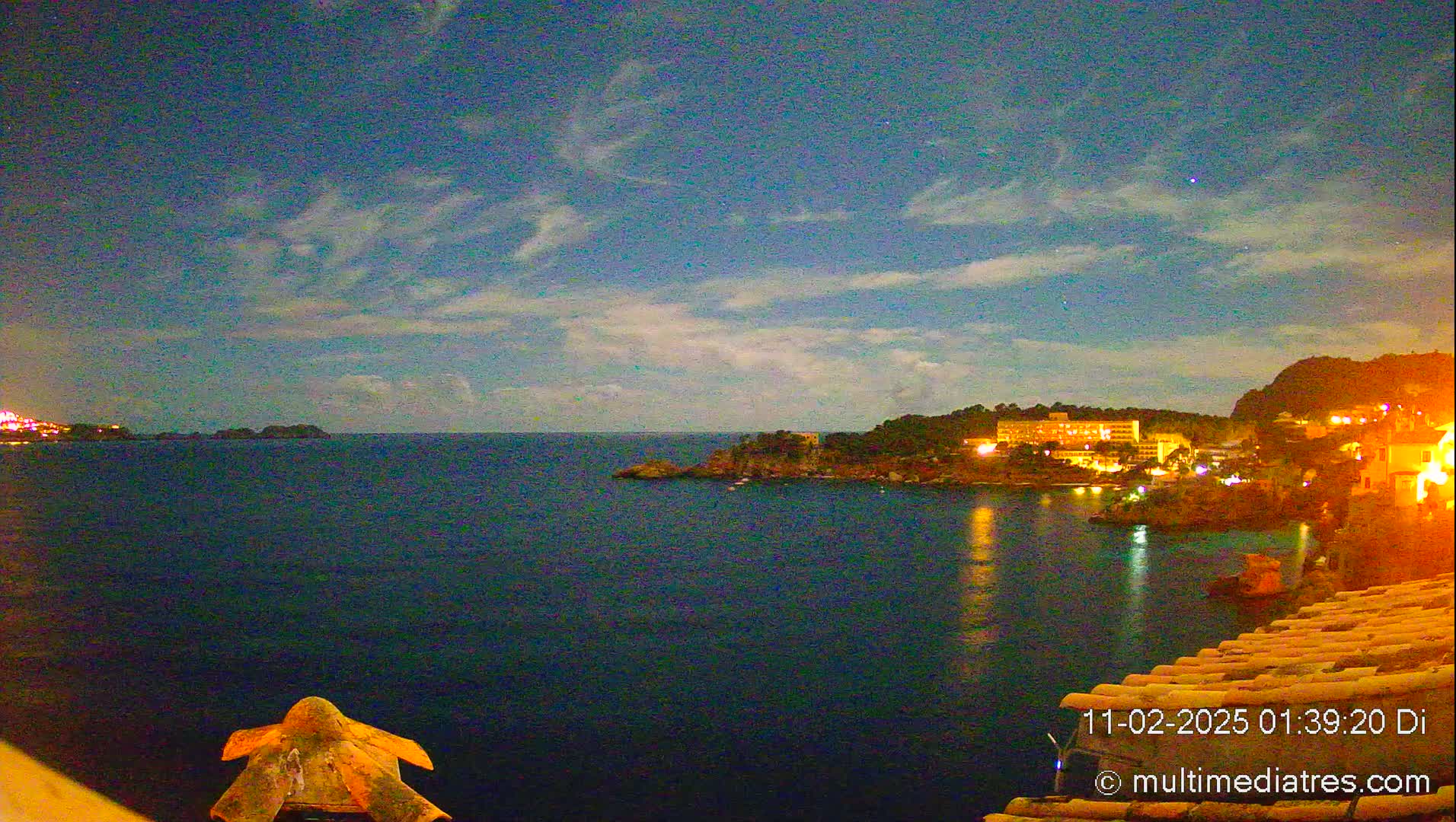 A nighttime view of a calm ocean reflecting the lights of a coastal town under a partly cloudy sky.