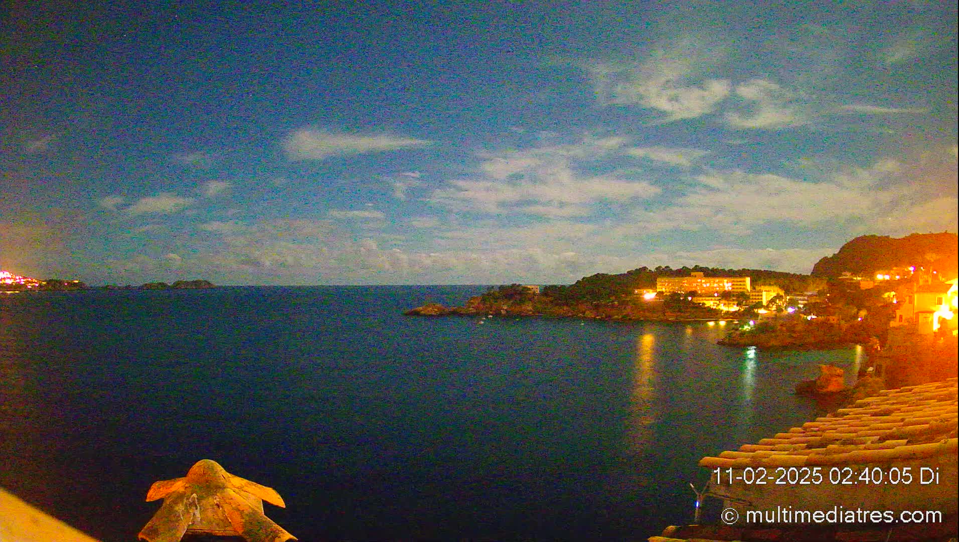 A nighttime coastal scene shows a dark blue sea reflecting the lights of a town nestled on a hill, under a mostly clear sky with scattered clouds.