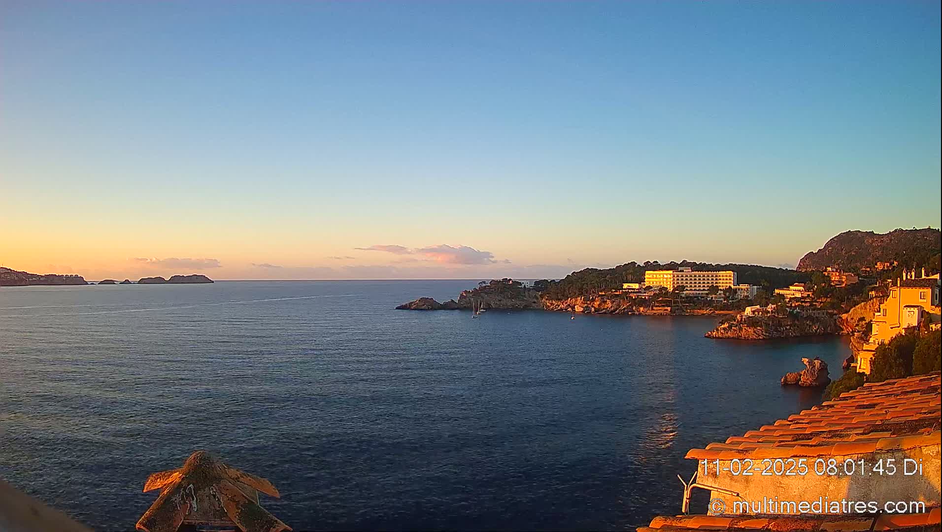 A calm ocean cove at sunrise, with a hotel and buildings nestled on the rocky coastline and islands in the distance.