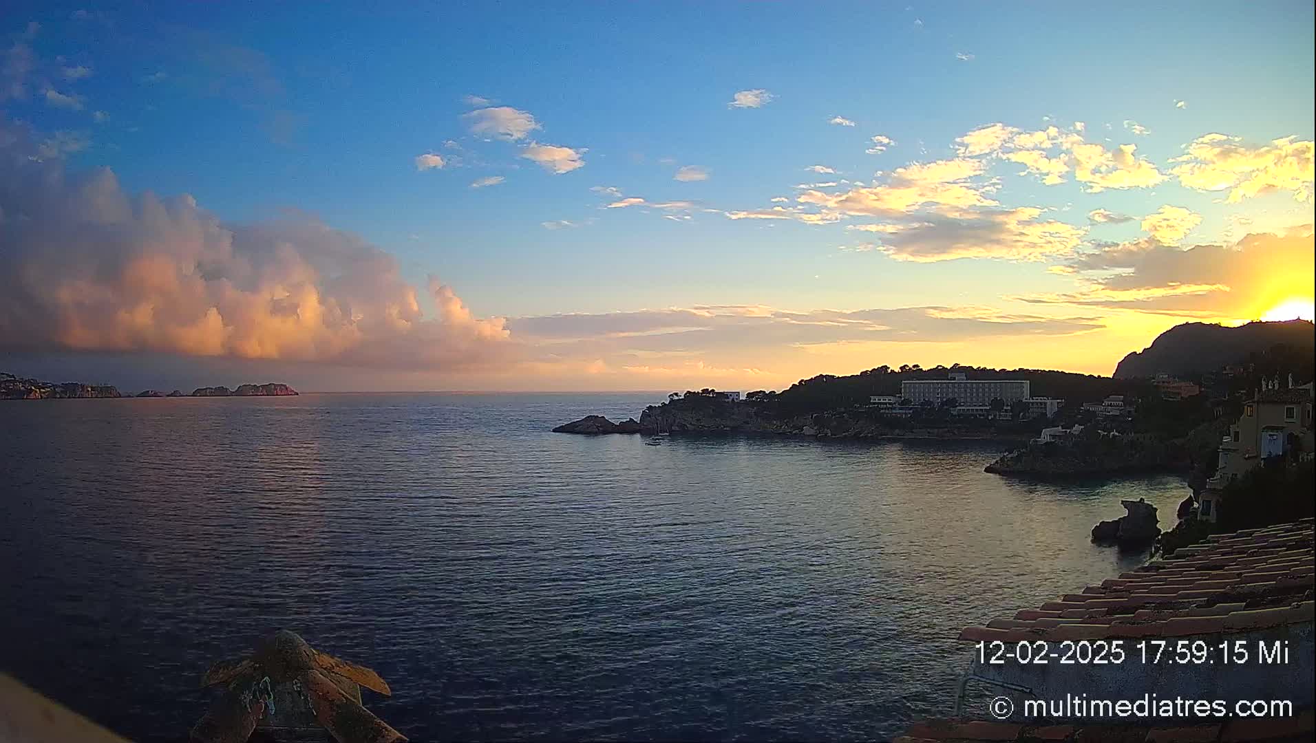 A coastal sunset scene shows a calm sea with rocky shores and buildings, under a partly cloudy sky with pink and orange hues.