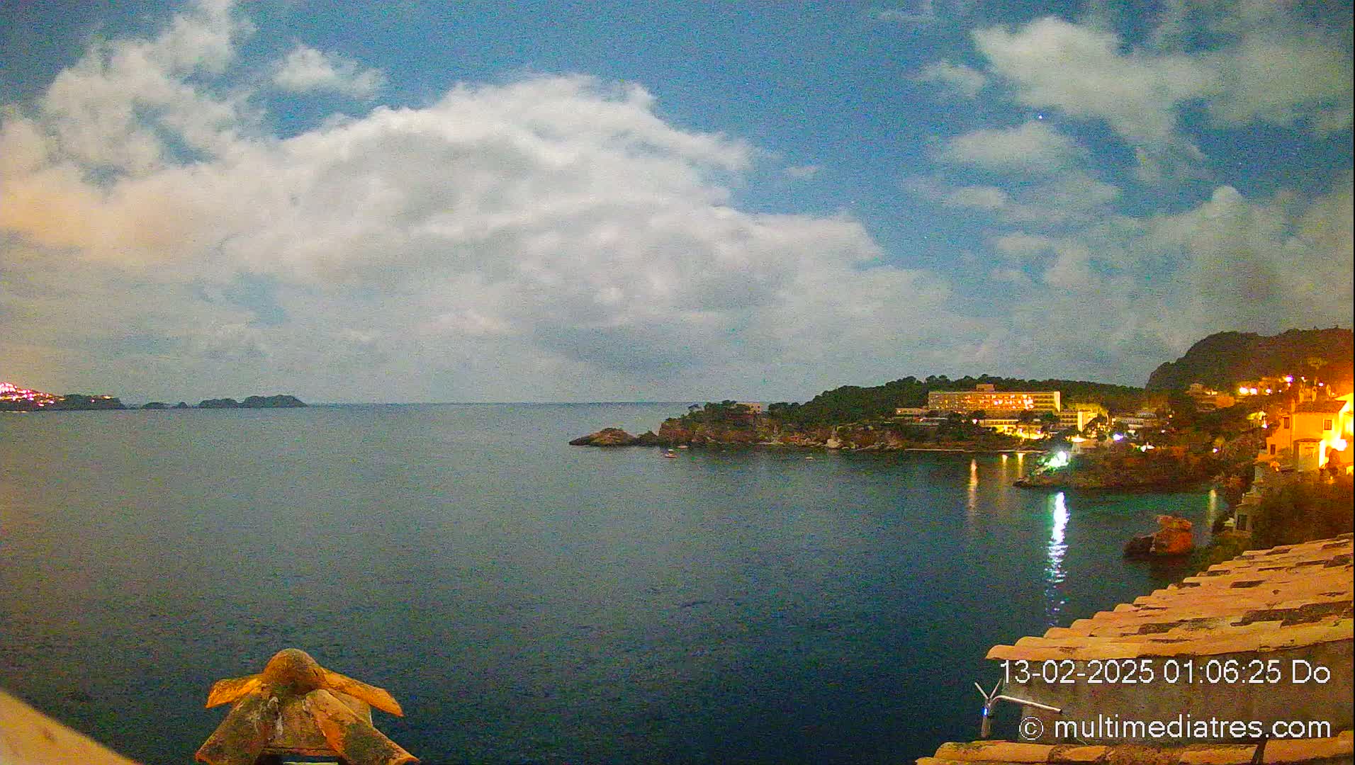 A nighttime coastal scene under a partly cloudy sky shows a calm body of water with a town nestled on a hillside and illuminated buildings along the shore.