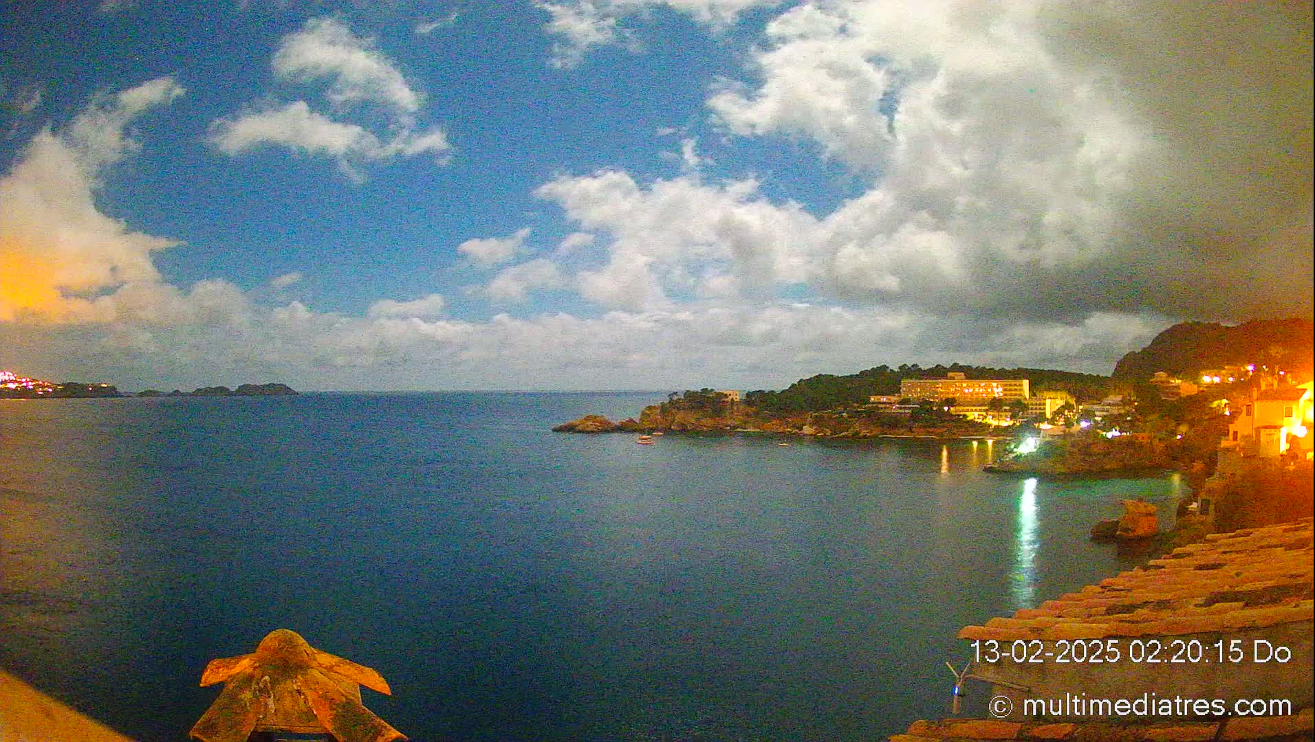 A nighttime coastal scene shows a calm, dark-blue sea under a partly cloudy sky, with a village of illuminated buildings nestled along a rocky shoreline.