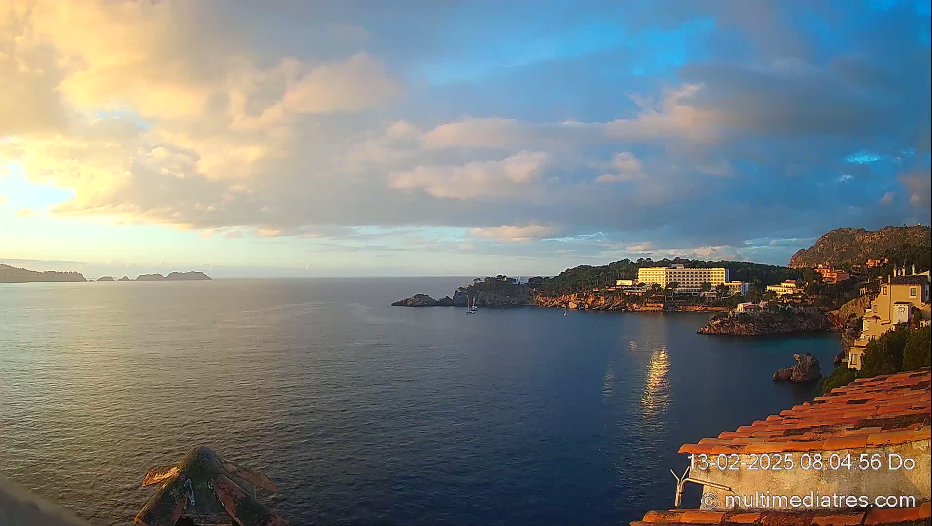 A coastal scene at sunrise shows a calm bay with a large hotel and other buildings nestled along a rocky shoreline, under a partly cloudy sky.