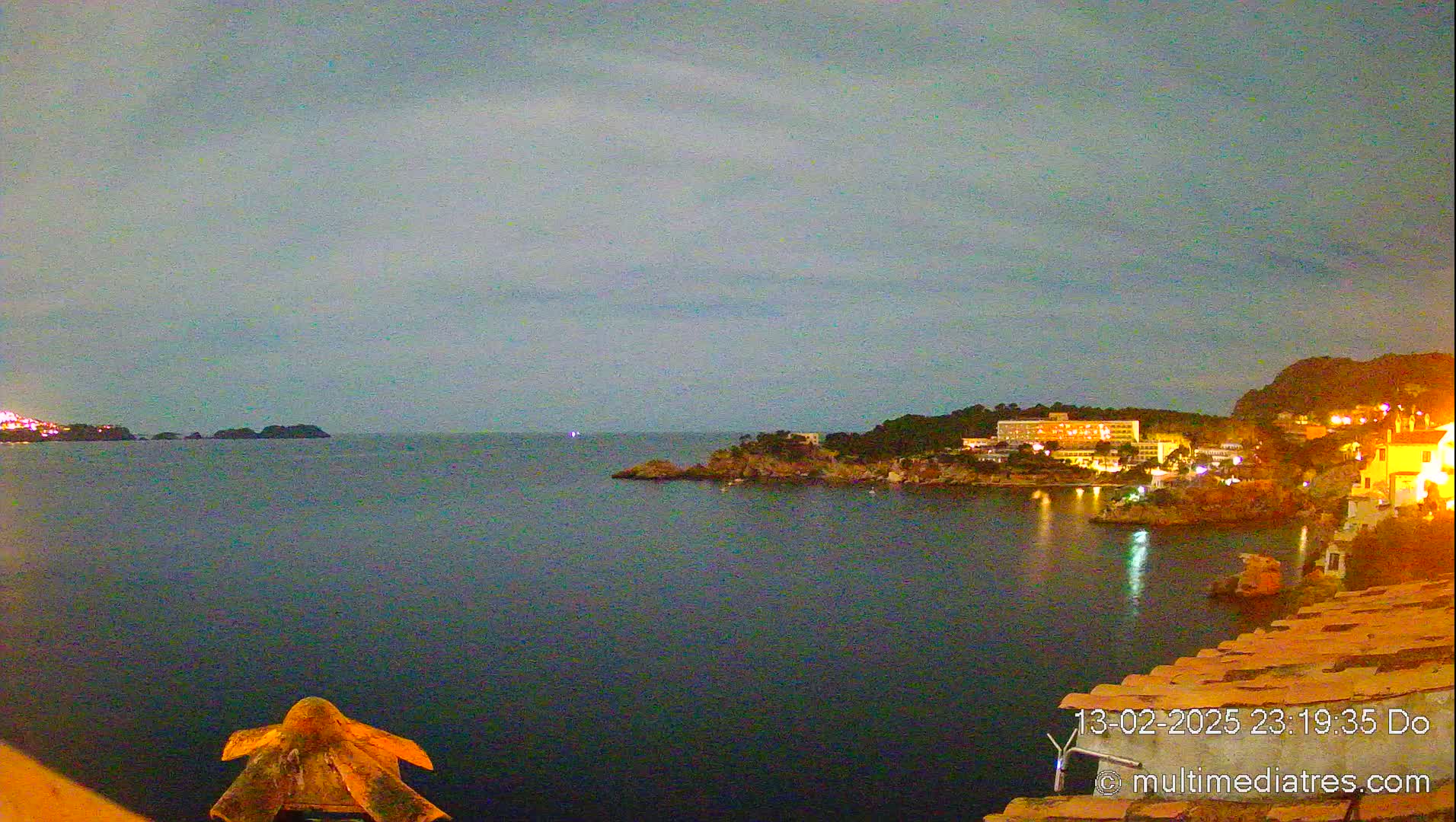 A nighttime view of a calm, dark-blue sea, with illuminated buildings and trees on a rocky coastline under a cloudy sky.