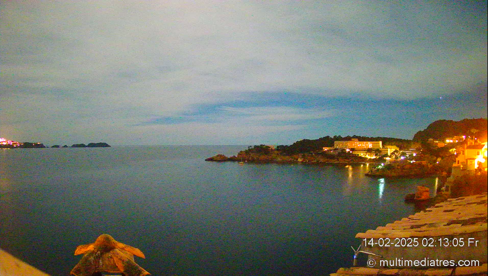 A nighttime view of a calm, dark-blue sea with a town's lights illuminating a rocky coastline under a twilight sky.