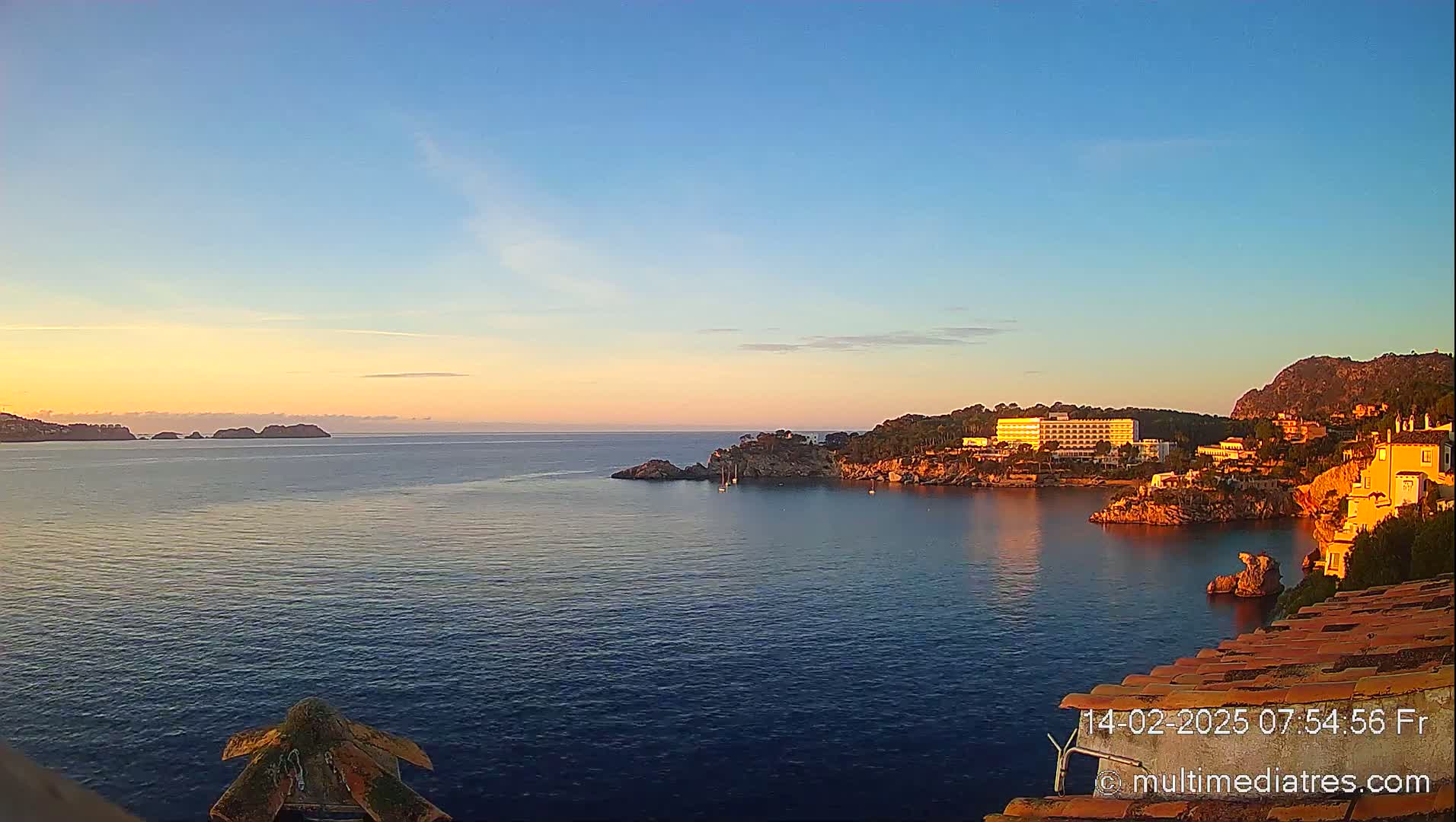 A calm, sunlit ocean bay at sunrise, featuring a coastal town nestled against a hillside and a few small islands in the distance.