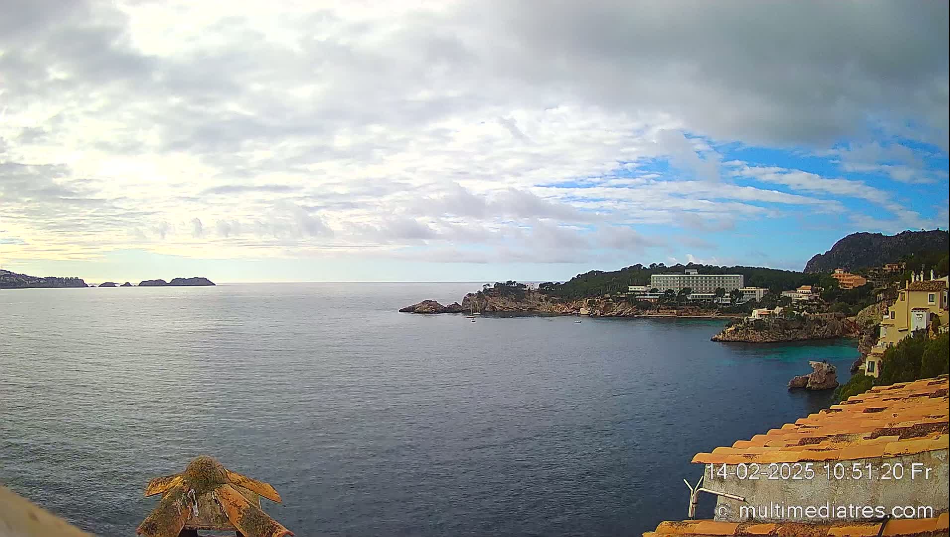 A partly cloudy day reveals a coastal scene with a calm sea, rocky headlands, and buildings nestled along the shoreline.