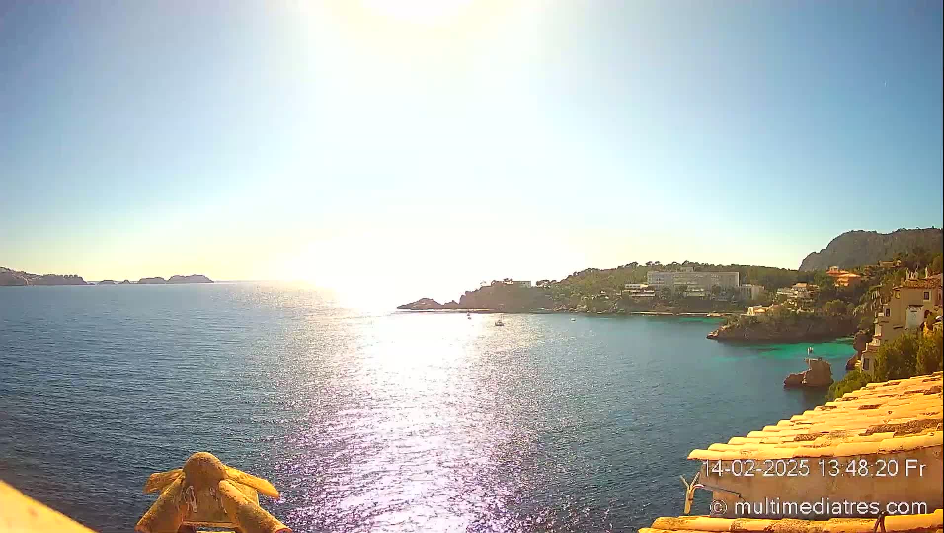 A sunny, bright day reveals a picturesque coastal scene with calm, sparkling water, buildings nestled into a rocky shoreline, and distant islands.