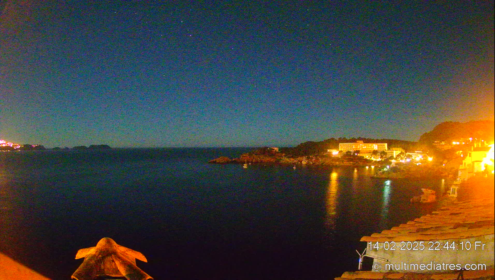 A nighttime coastal scene shows a dark body of water reflecting the lights of buildings on a hillside under a clear, starry sky.