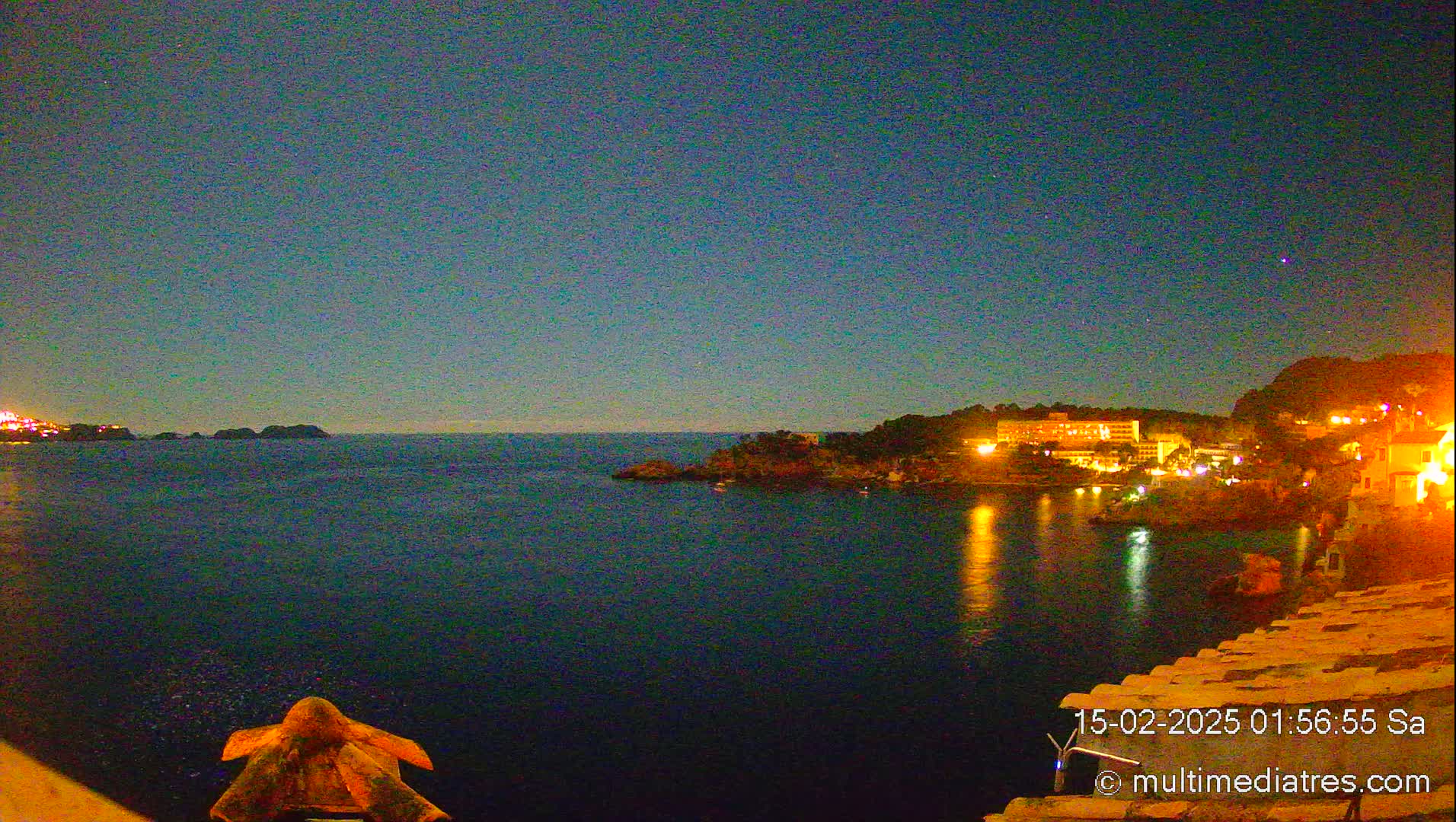 A nighttime coastal scene shows a dark body of water reflecting the lights of buildings on a nearby shore under a clear, dark sky.