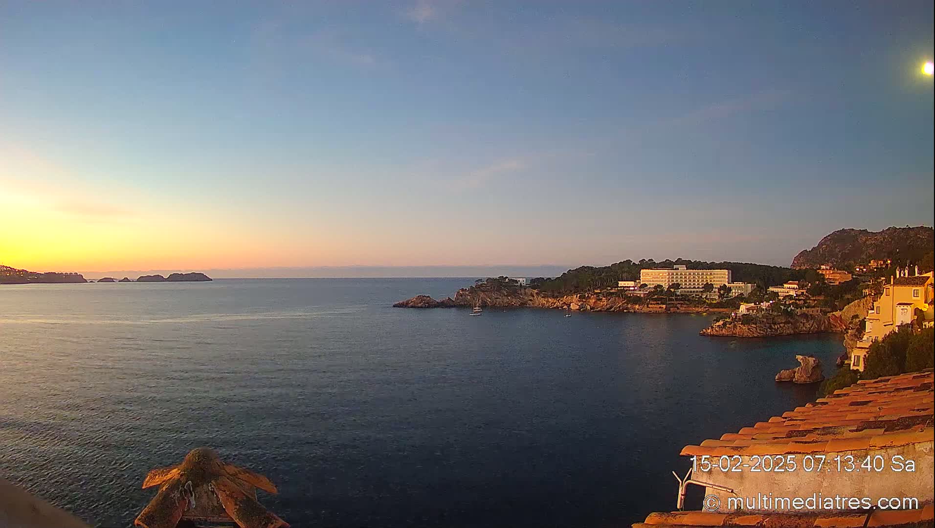 A calm ocean at sunset, with a large hotel and other buildings situated on a rocky coast, and small islands in the distance.