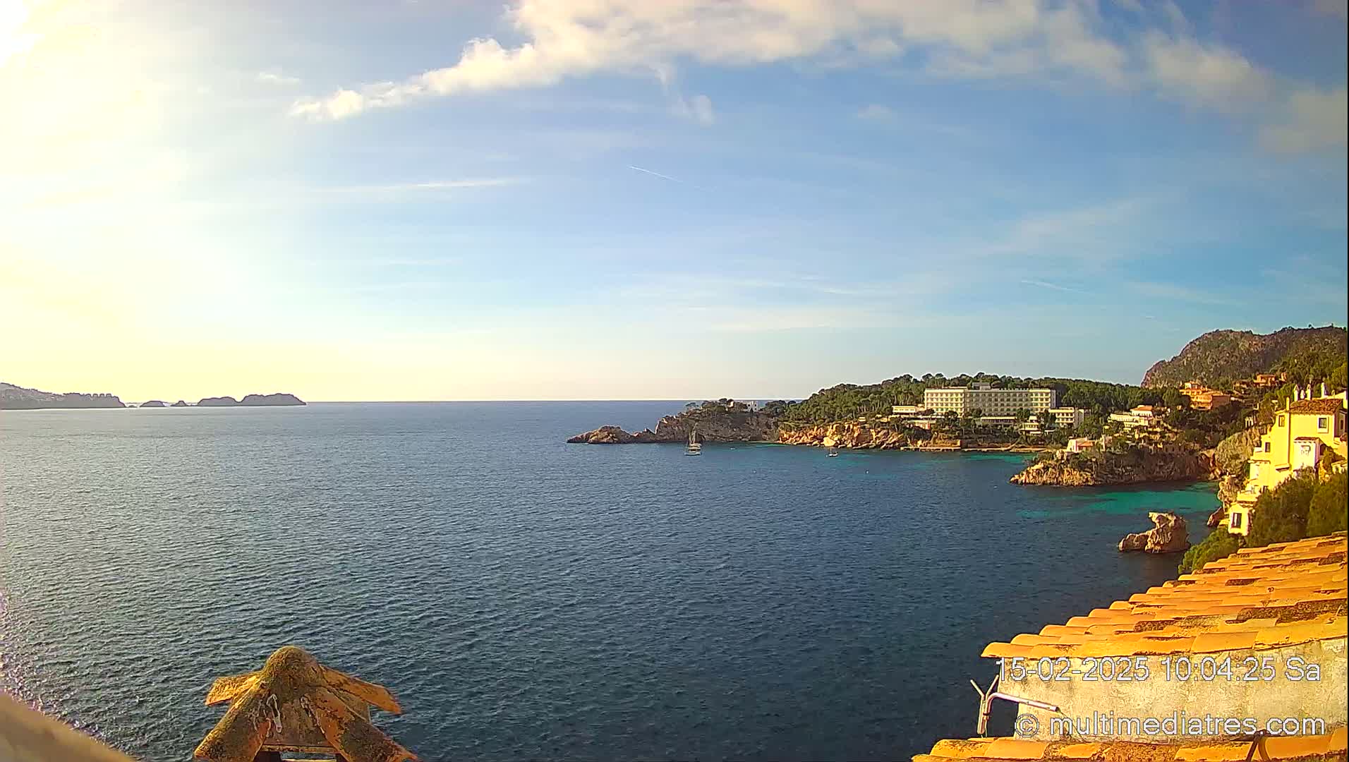A partly sunny day reveals a coastal scene of a calm blue sea with a hotel and buildings situated on a rocky, tree-covered hillside.