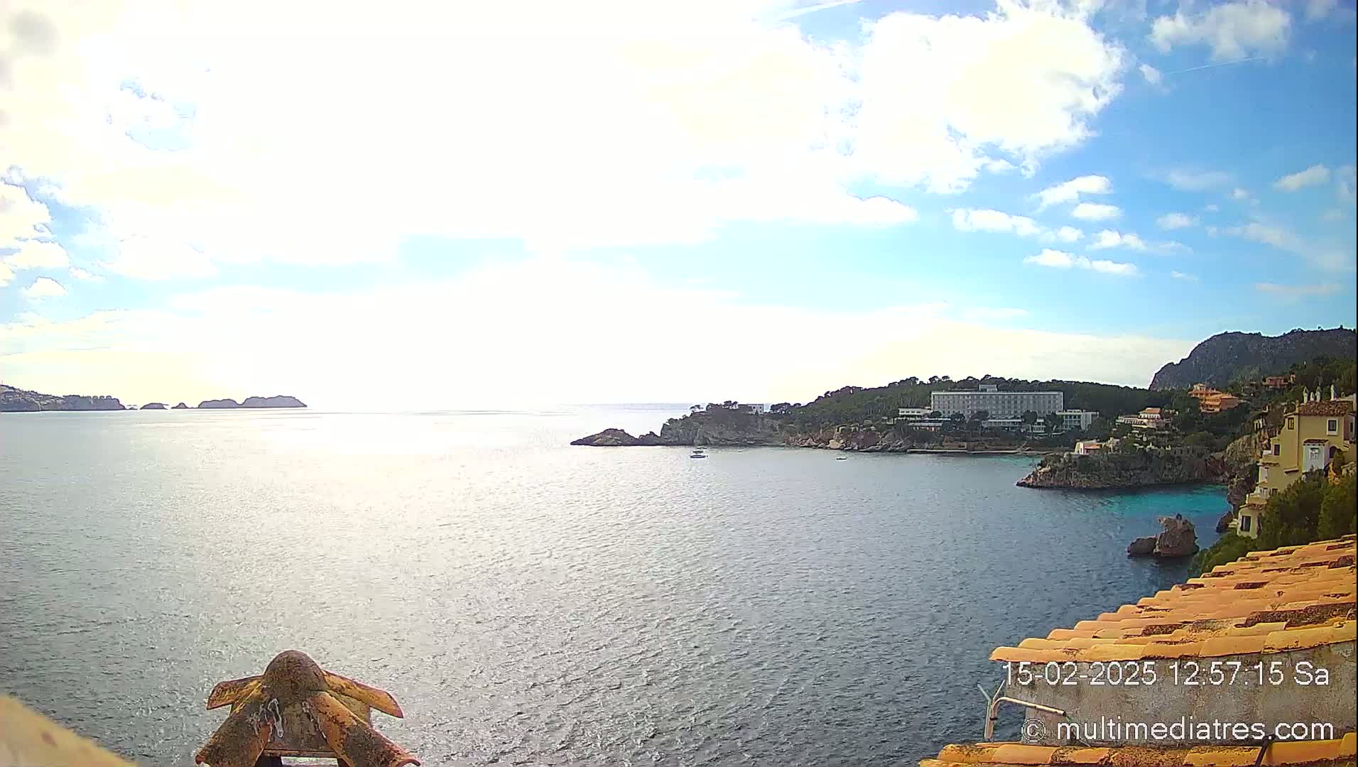 A partly cloudy day shows a coastal view of a calm sea with small islands, a hotel, and buildings along a rocky shoreline.