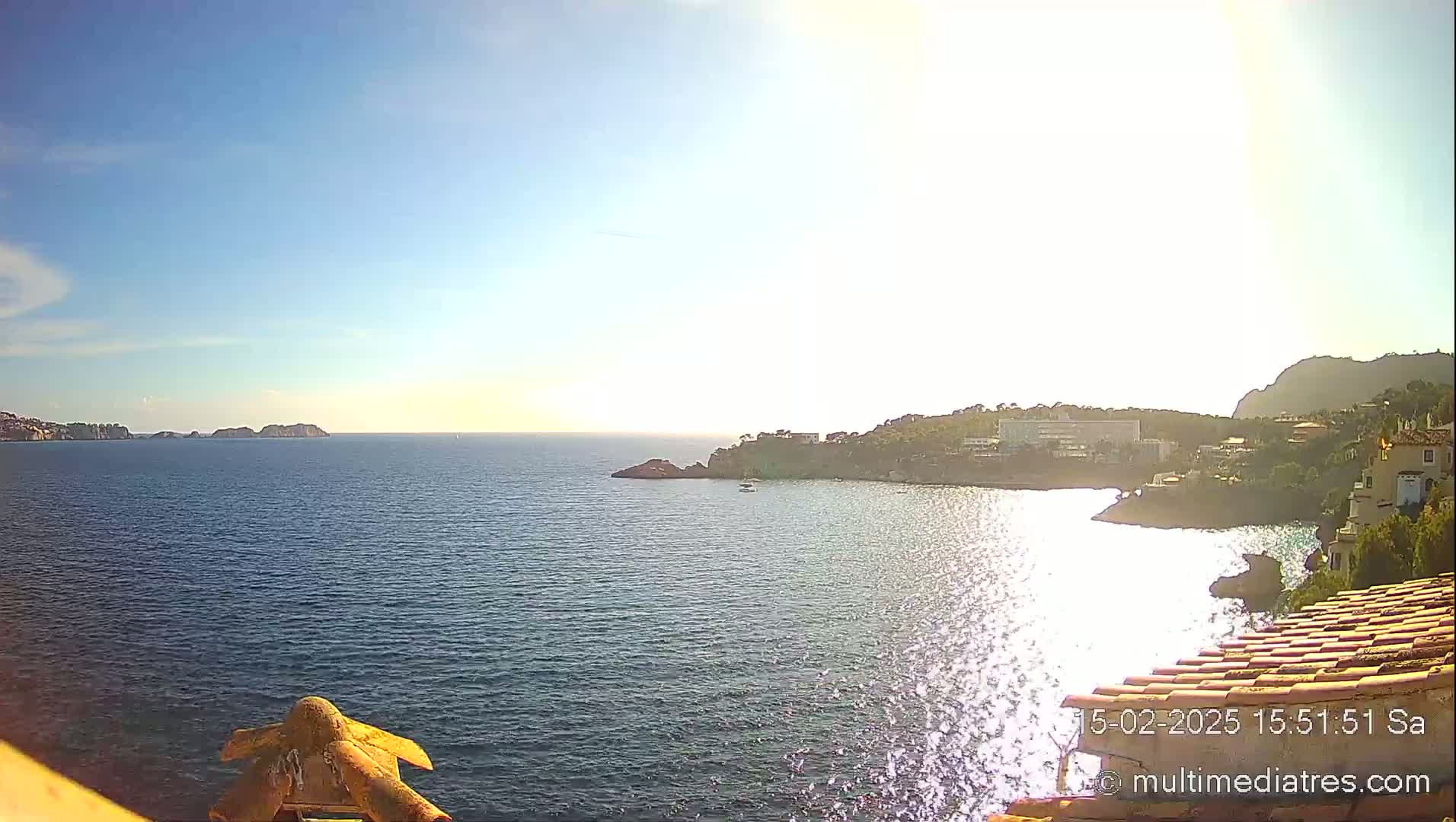 A sunny, calm ocean bay is viewed from a high vantage point, showing a coastline with buildings and distant islands.