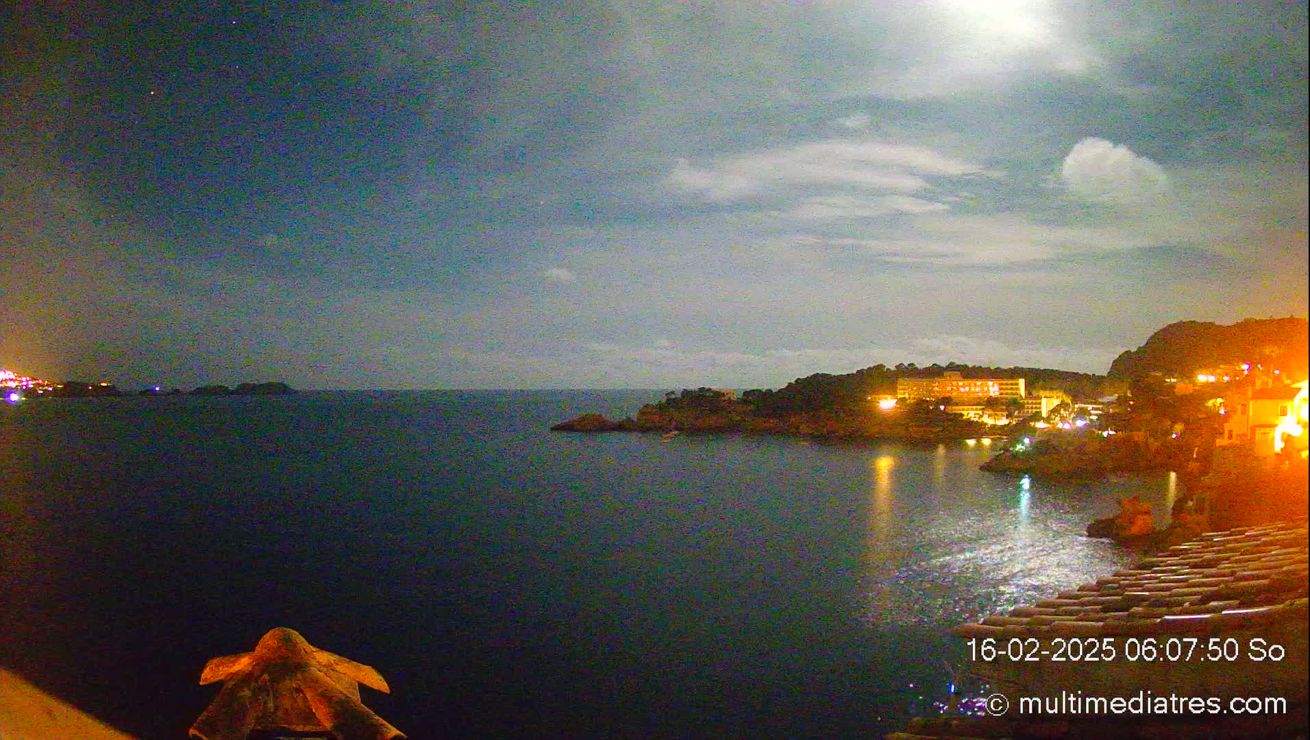 A nighttime view of a calm, dark sea under a partly cloudy moonlit sky shows a coastline with buildings lit up.