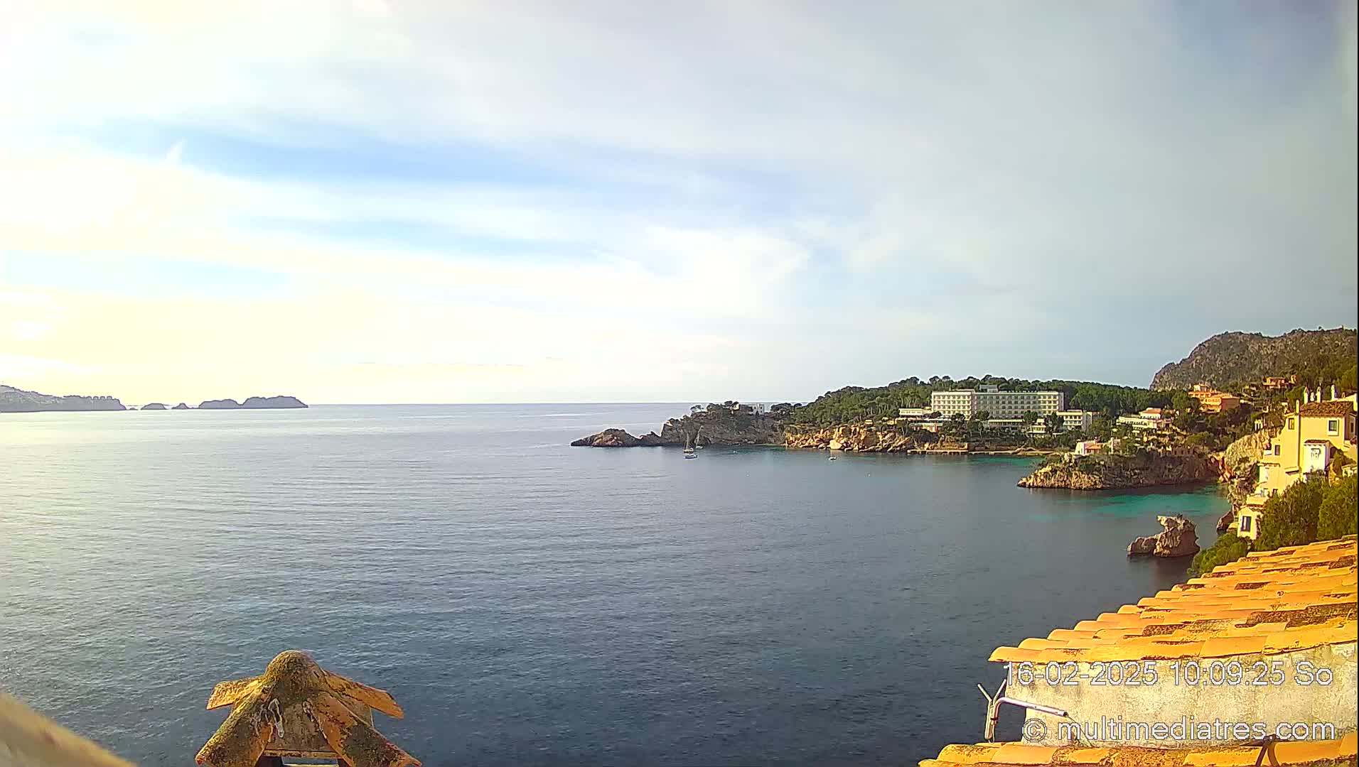 A tranquil coastal scene under a partly cloudy sky shows a calm sea with a shoreline featuring buildings and rocky outcrops, and distant islands.