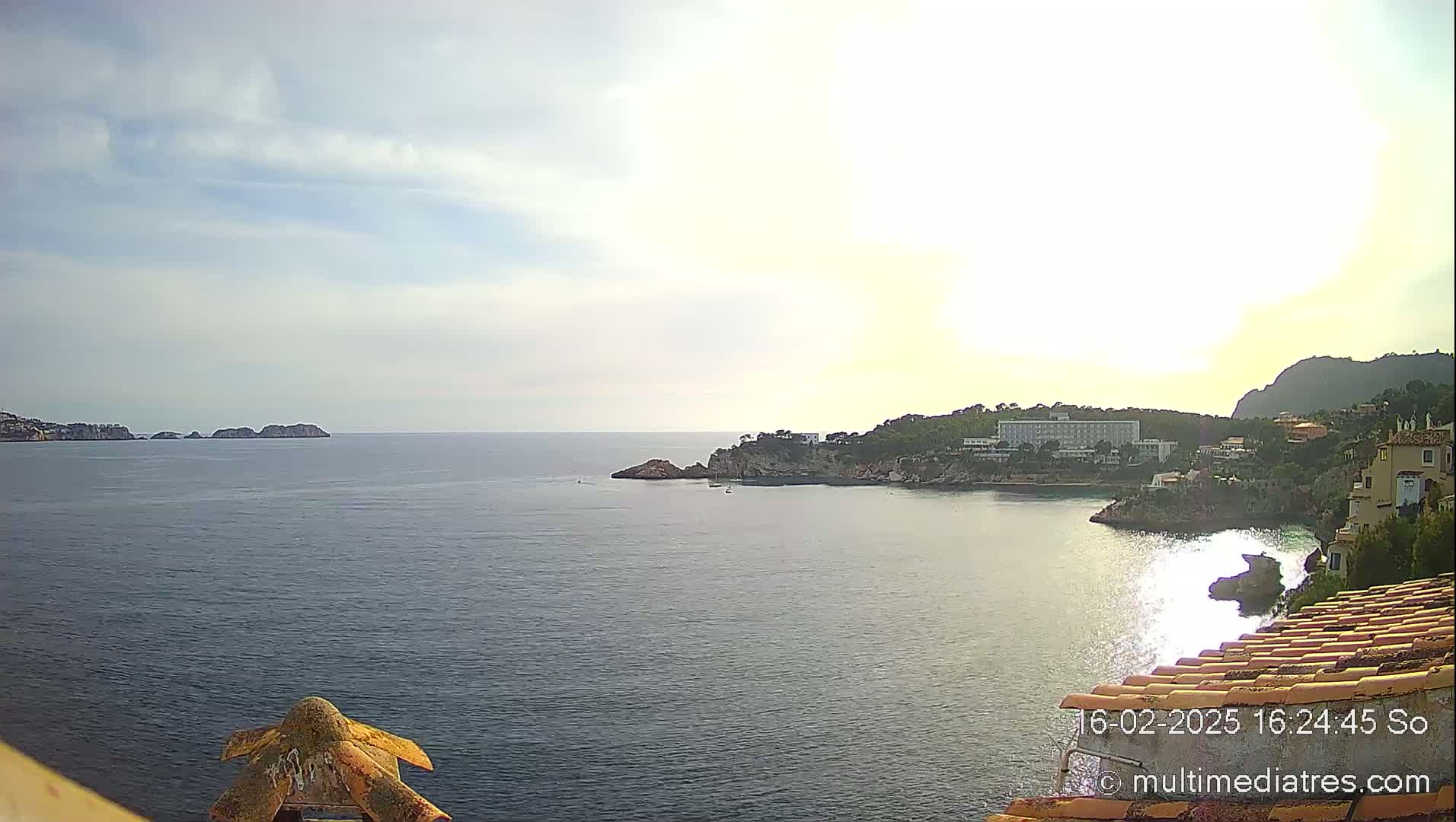 A partly cloudy day reveals a calm sea with a rocky coastline featuring buildings and a larger hotel, as well as distant islands.