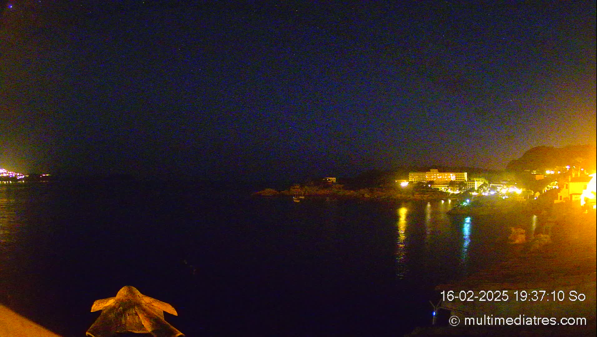 A nighttime view of a calm body of water reflecting the lights of a town on the opposite shore under a clear, starry sky.