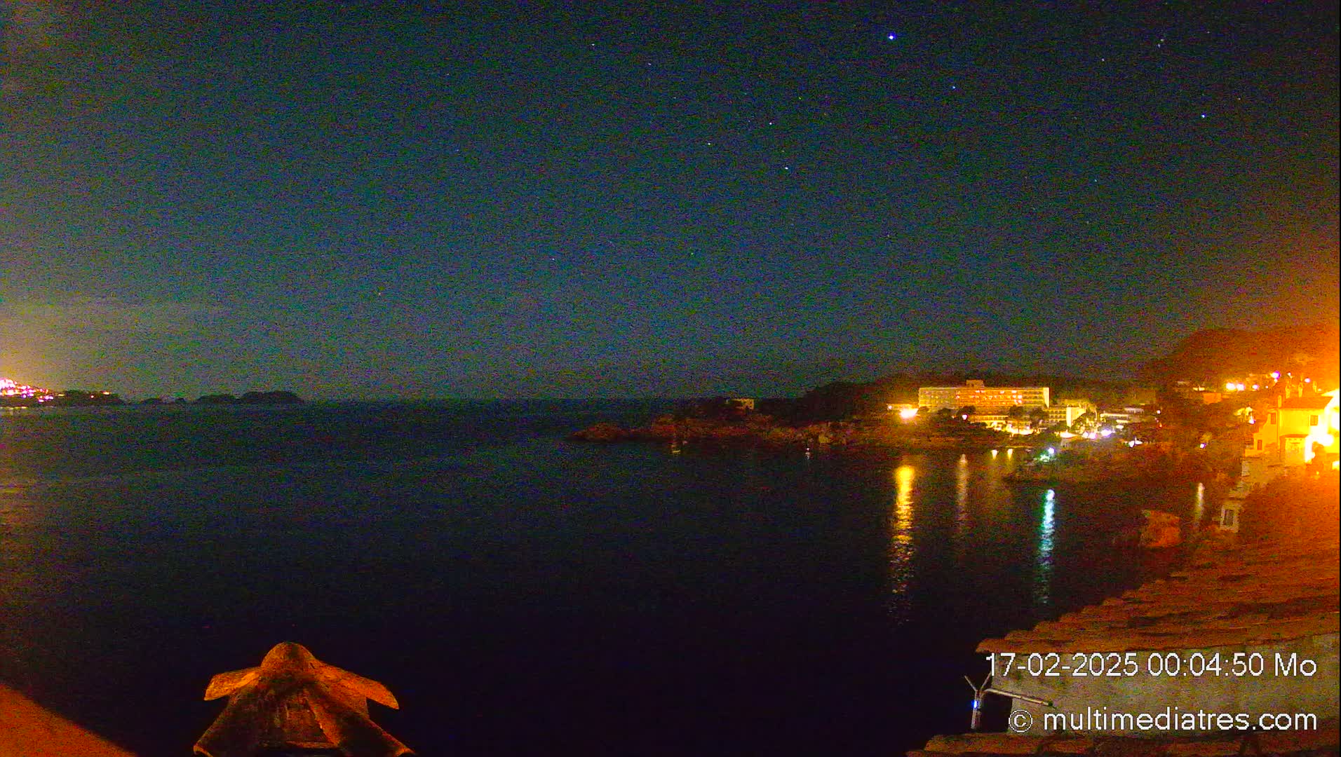A nighttime coastal scene under a clear, starry sky shows a calm body of water reflecting the lights of buildings on the shore.