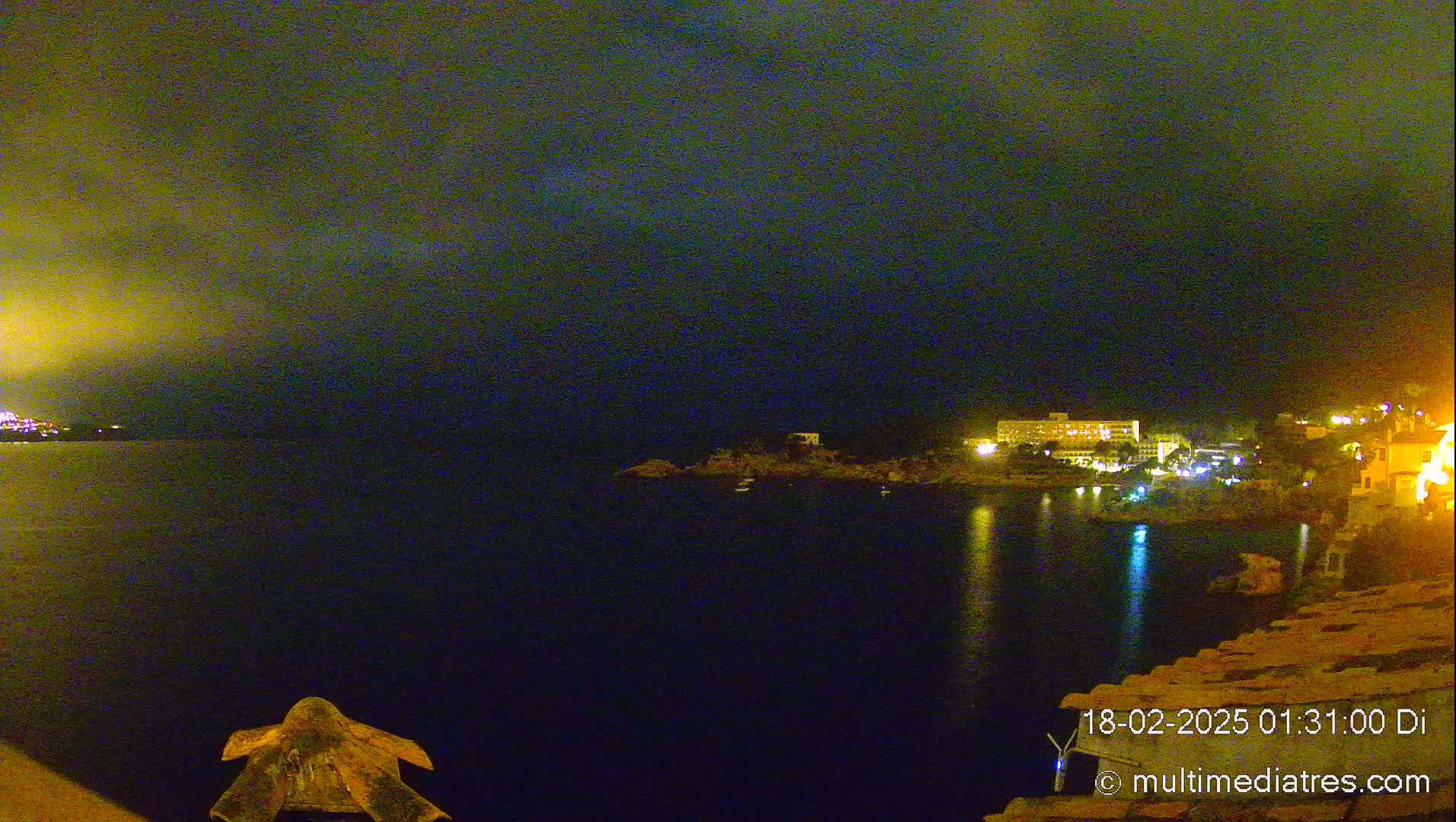 A nighttime view of a dark body of water reflecting lights from buildings on the opposite shore under a cloudy sky.