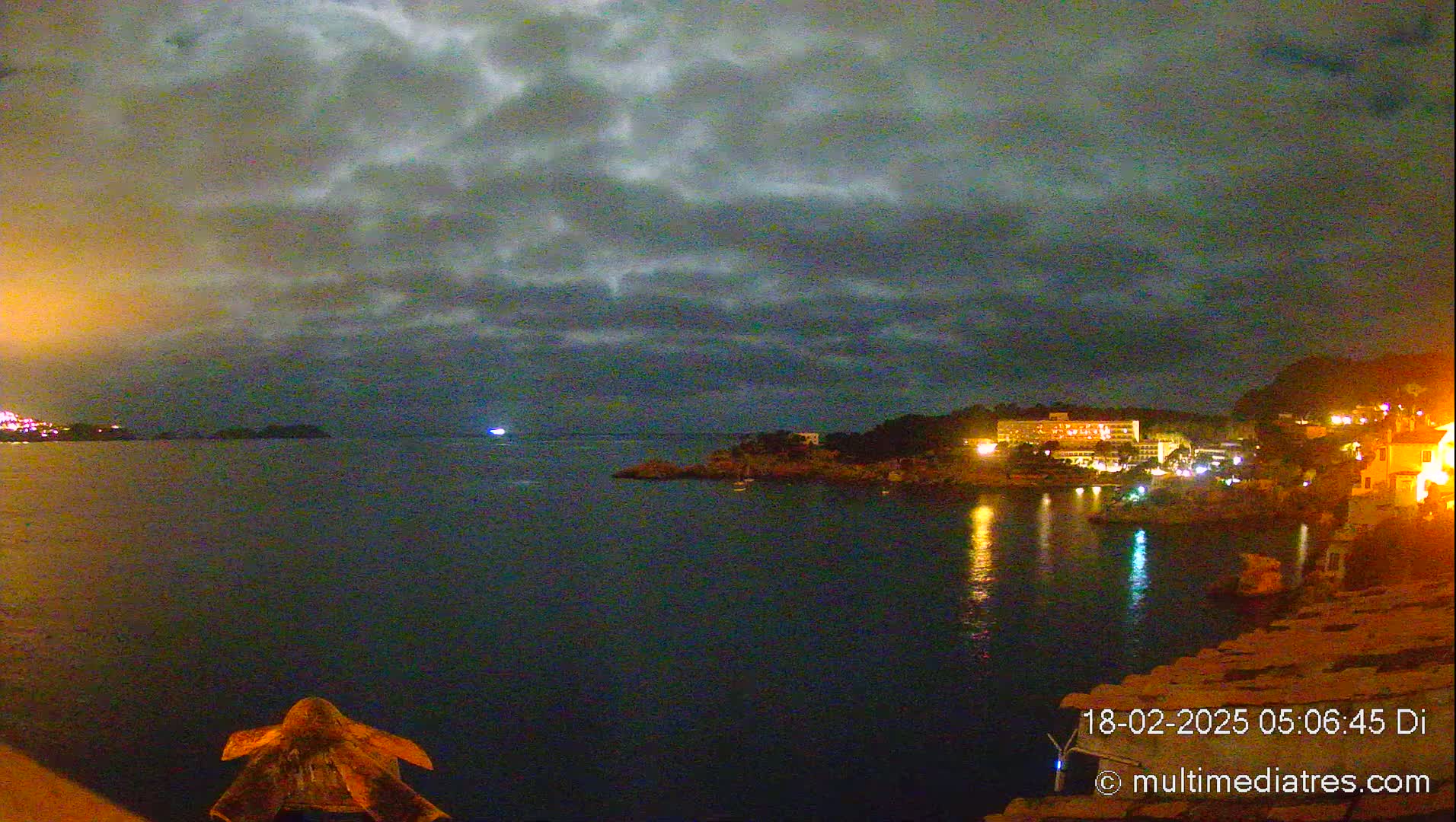 A nighttime coastal scene under a cloudy sky shows a dark body of water reflecting the lights of buildings on the shore.